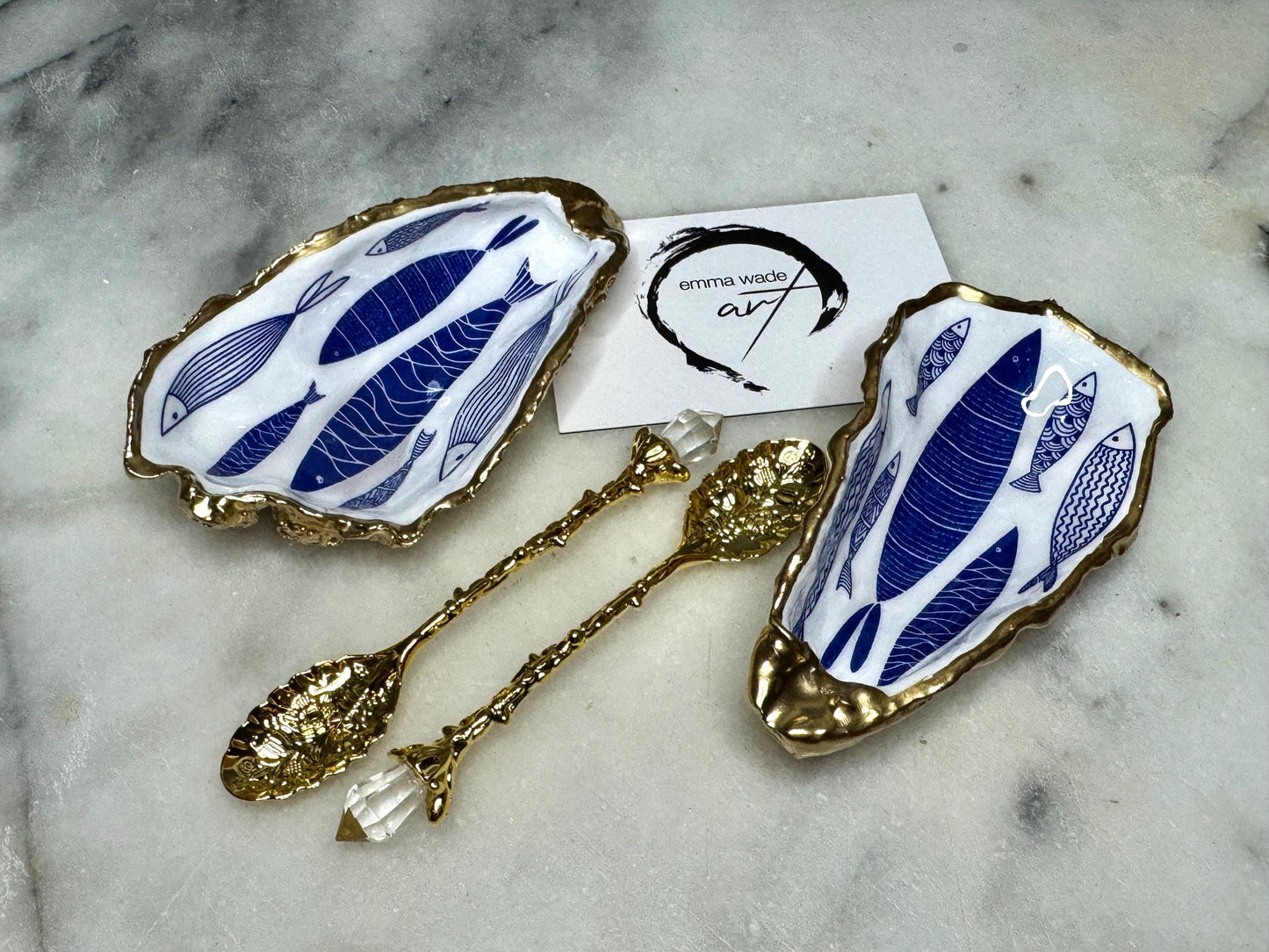 Pair of handmade Dorset oyster shell salt and pepper pinch pots decorated with elegant blue and white fish artwork, finished with hand applied liquid gold leaf edging and sealed in glossy food-safe resin. Coastal shell cruet dishes styled with optional gold crystal spoons on a marble background. Luxury nautical kitchen decor inspired by the sea, perfect for beach house dining, dinner parties, weddings, anniversaries, and coastal home gifts.
