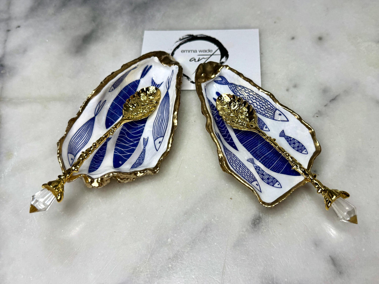 Pair of handmade Dorset oyster shell salt and pepper pinch pots decorated with elegant blue and white fish artwork, finished with hand applied liquid gold leaf edging and sealed in glossy food-safe resin. Coastal shell cruet dishes styled with optional gold crystal spoons on a marble background. Luxury nautical kitchen decor inspired by the sea, perfect for beach house dining, dinner parties, weddings, anniversaries, and coastal home gifts.