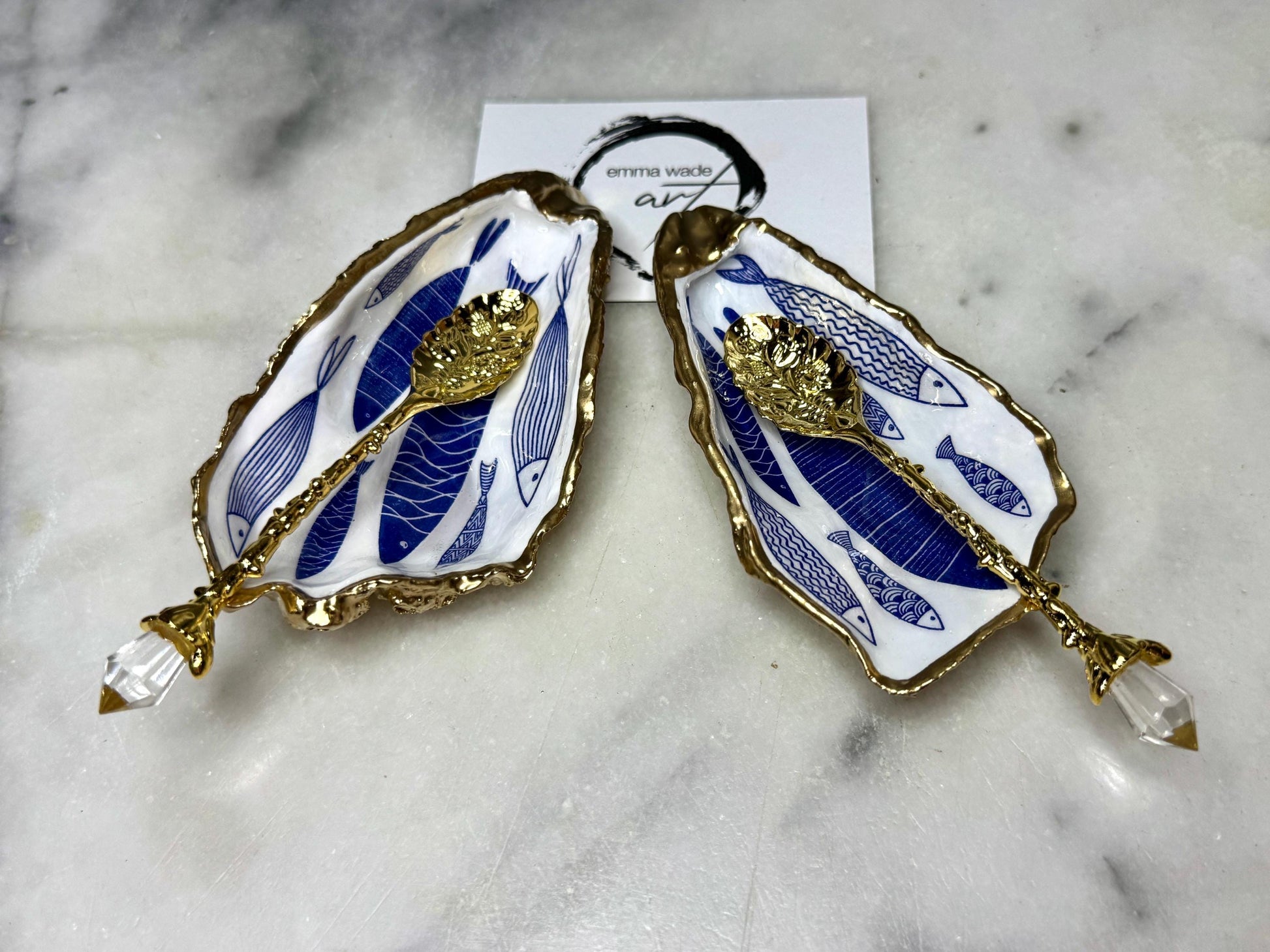Pair of handmade Dorset oyster shell salt and pepper pinch pots decorated with elegant blue and white fish artwork, finished with hand applied liquid gold leaf edging and sealed in glossy food-safe resin. Coastal shell cruet dishes styled with optional gold crystal spoons on a marble background. Luxury nautical kitchen decor inspired by the sea, perfect for beach house dining, dinner parties, weddings, anniversaries, and coastal home gifts.