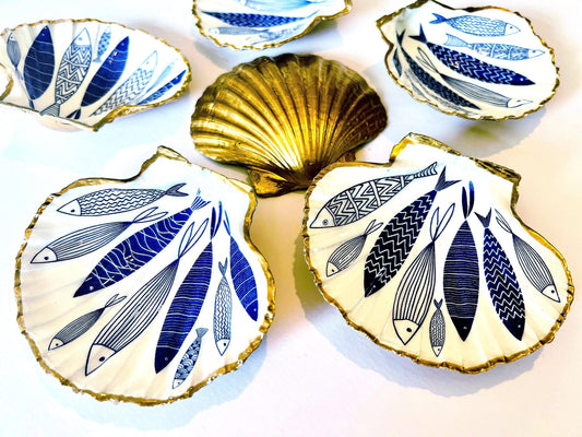 Handmade Dorset scallop shell trinket dishes featuring elegant blue fish illustrations with hand-painted gold leaf edging and glossy crystal clear resin. Coastal nautical jewellery dishes inspired by Scandinavian ocean decor, styled on a neutral background with detailed blue fish artwork and luxurious gilded edges.