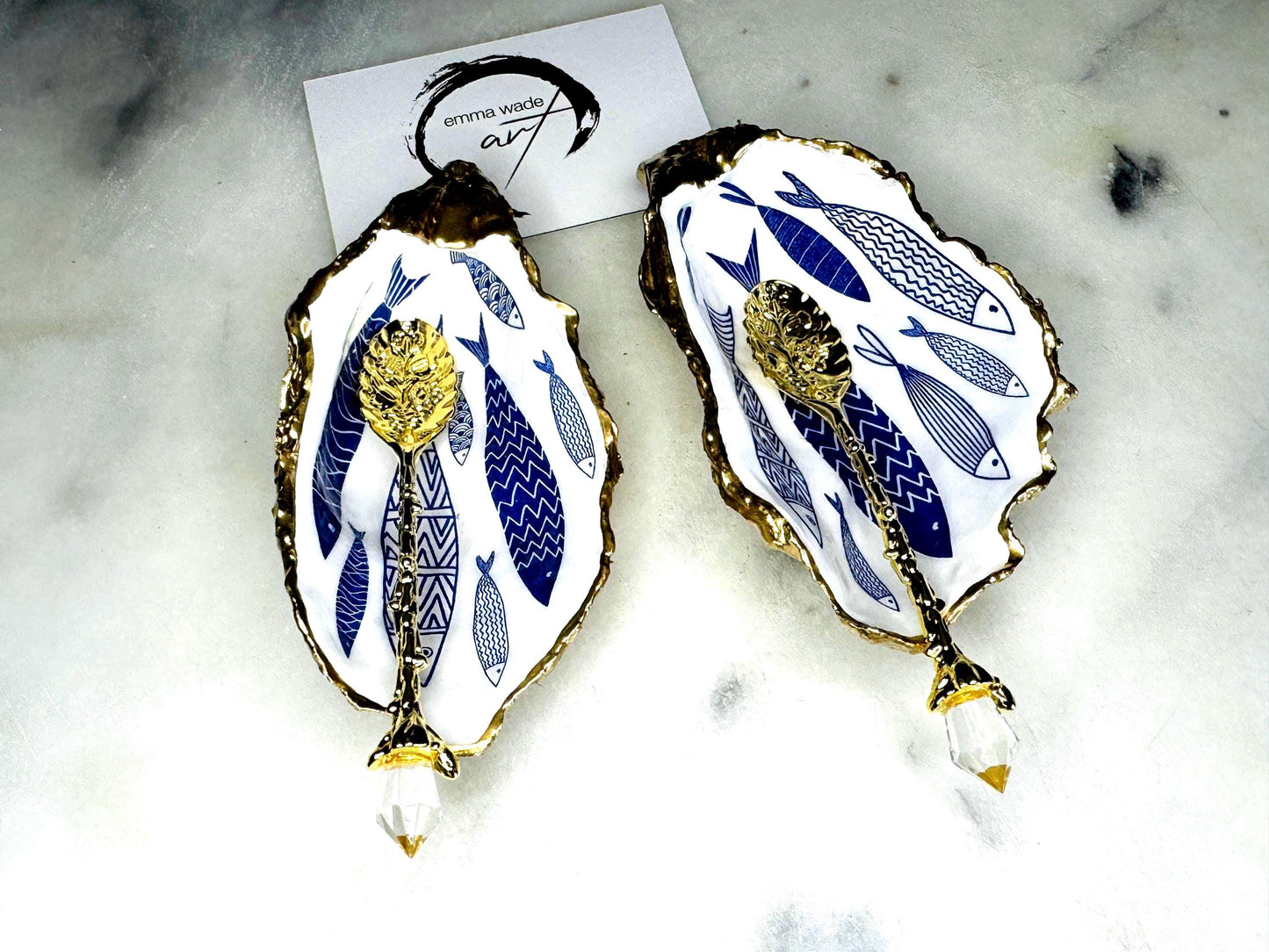 Pair of handmade Dorset oyster shell salt and pepper pinch pots decorated with elegant blue and white fish artwork, finished with hand applied liquid gold leaf edging and sealed in glossy food-safe resin. Coastal shell cruet dishes styled with optional gold crystal spoons on a marble background. Luxury nautical kitchen decor inspired by the sea, perfect for beach house dining, dinner parties, weddings, anniversaries, and coastal home gifts.