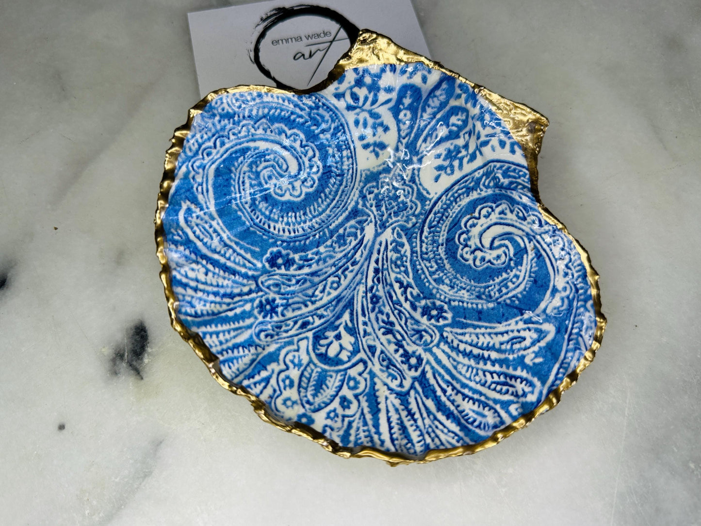 Blue and white paisley scallop shell ring catcher with hand applied gold leaf edging and glossy resin finish. Handmade Dorset shell jewellery tray styled with pearls on a marble background. Coastal vanity decor and luxury beach house accessory gift.