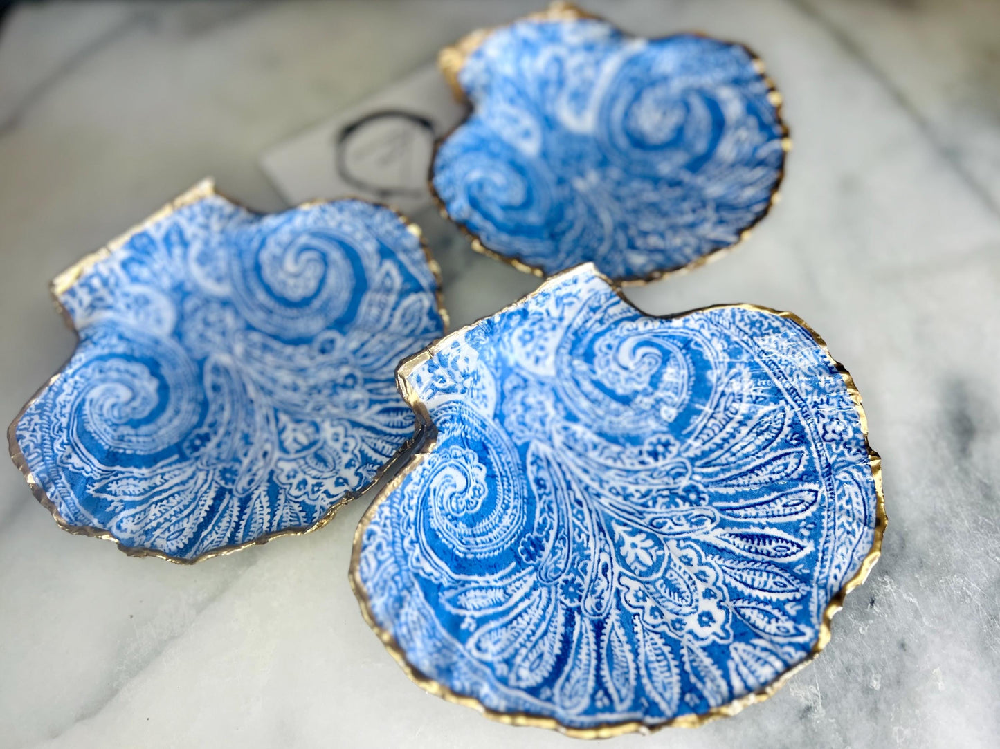 Blue and white paisley scallop shell ring catcher with hand applied gold leaf edging and glossy resin finish. Handmade Dorset shell jewellery tray styled with pearls on a marble background. Coastal vanity decor and luxury beach house accessory gift.