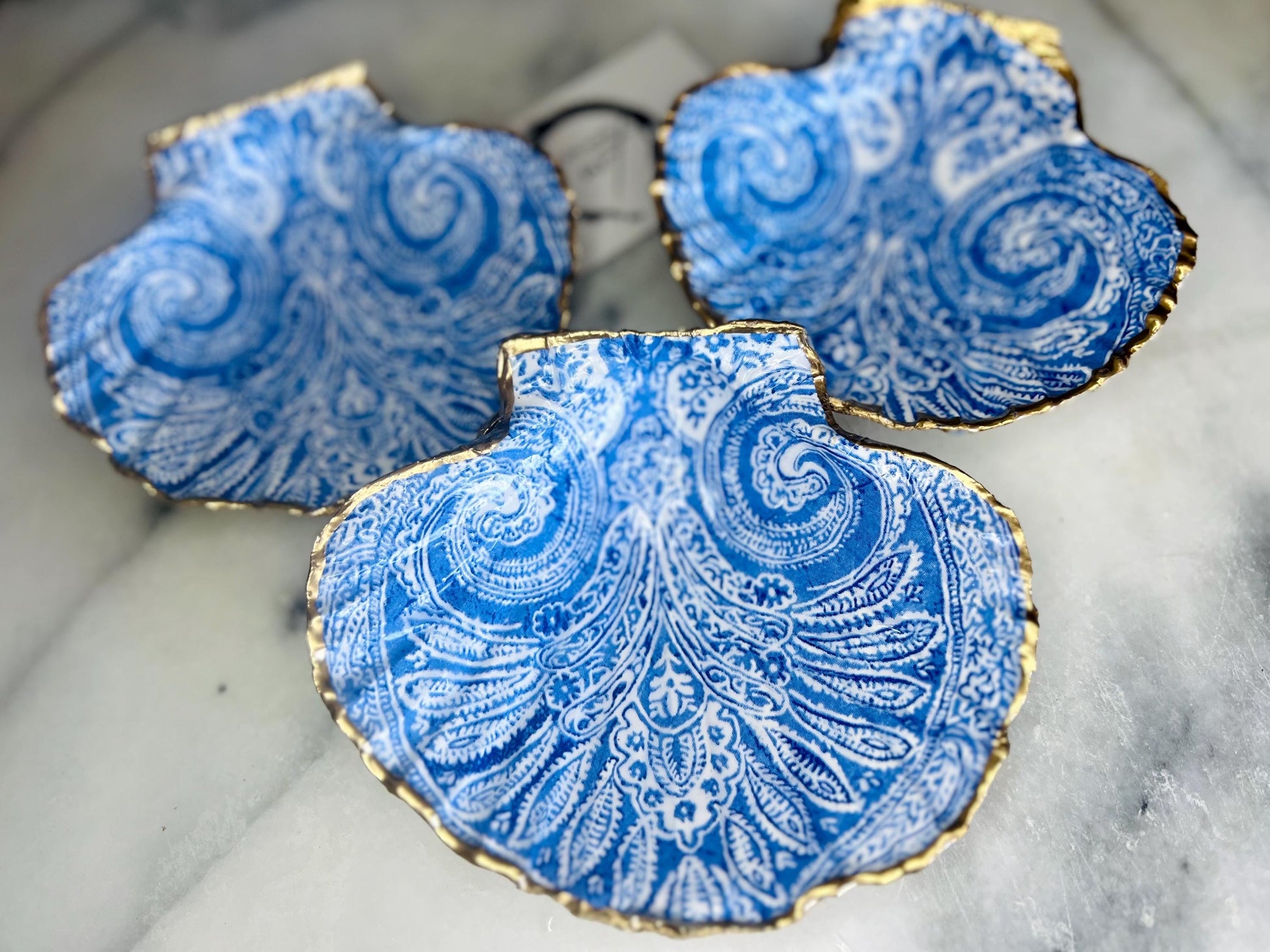 Blue and white paisley scallop shell ring catcher with hand applied gold leaf edging and glossy resin finish. Handmade Dorset shell jewellery tray styled with pearls on a marble background. Coastal vanity decor and luxury beach house accessory gift.
