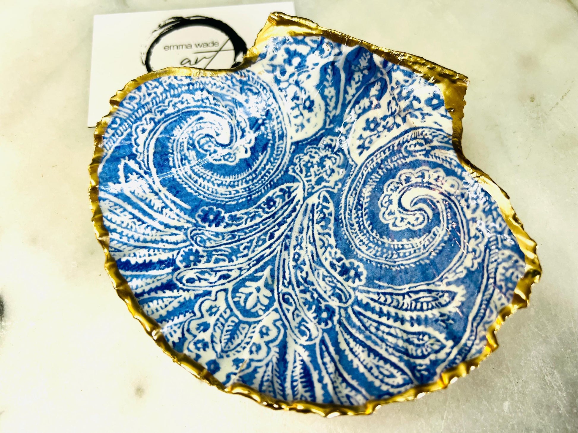 Blue and white paisley scallop shell ring catcher with hand applied gold leaf edging and glossy resin finish. Handmade Dorset shell jewellery tray styled with pearls on a marble background. Coastal vanity decor and luxury beach house accessory gift.