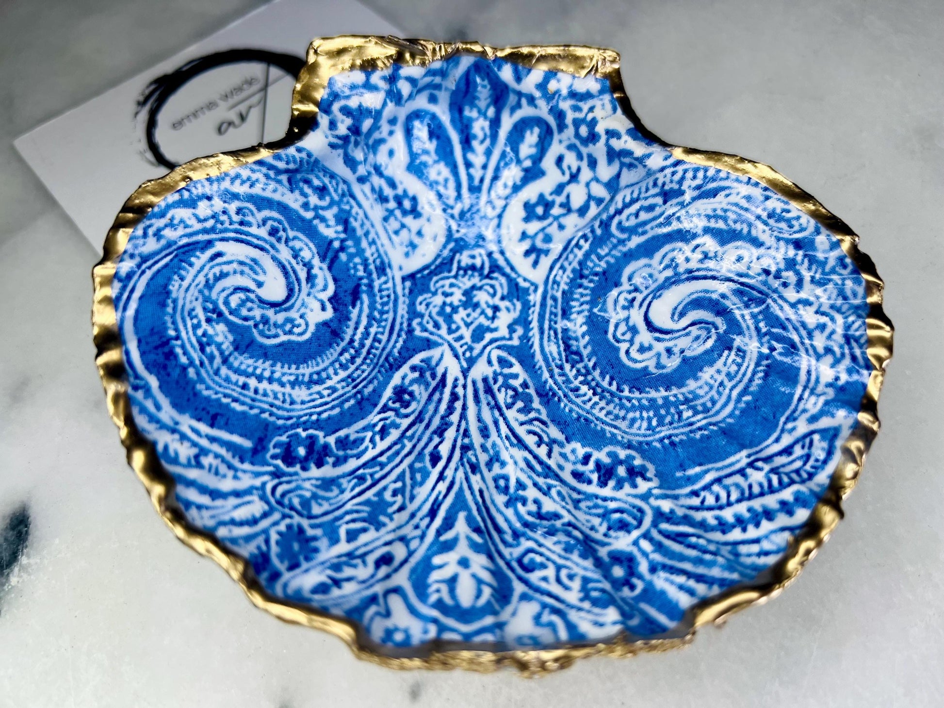 Blue and white paisley scallop shell ring catcher with hand applied gold leaf edging and glossy resin finish. Handmade Dorset shell jewellery tray styled with pearls on a marble background. Coastal vanity decor and luxury beach house accessory gift.