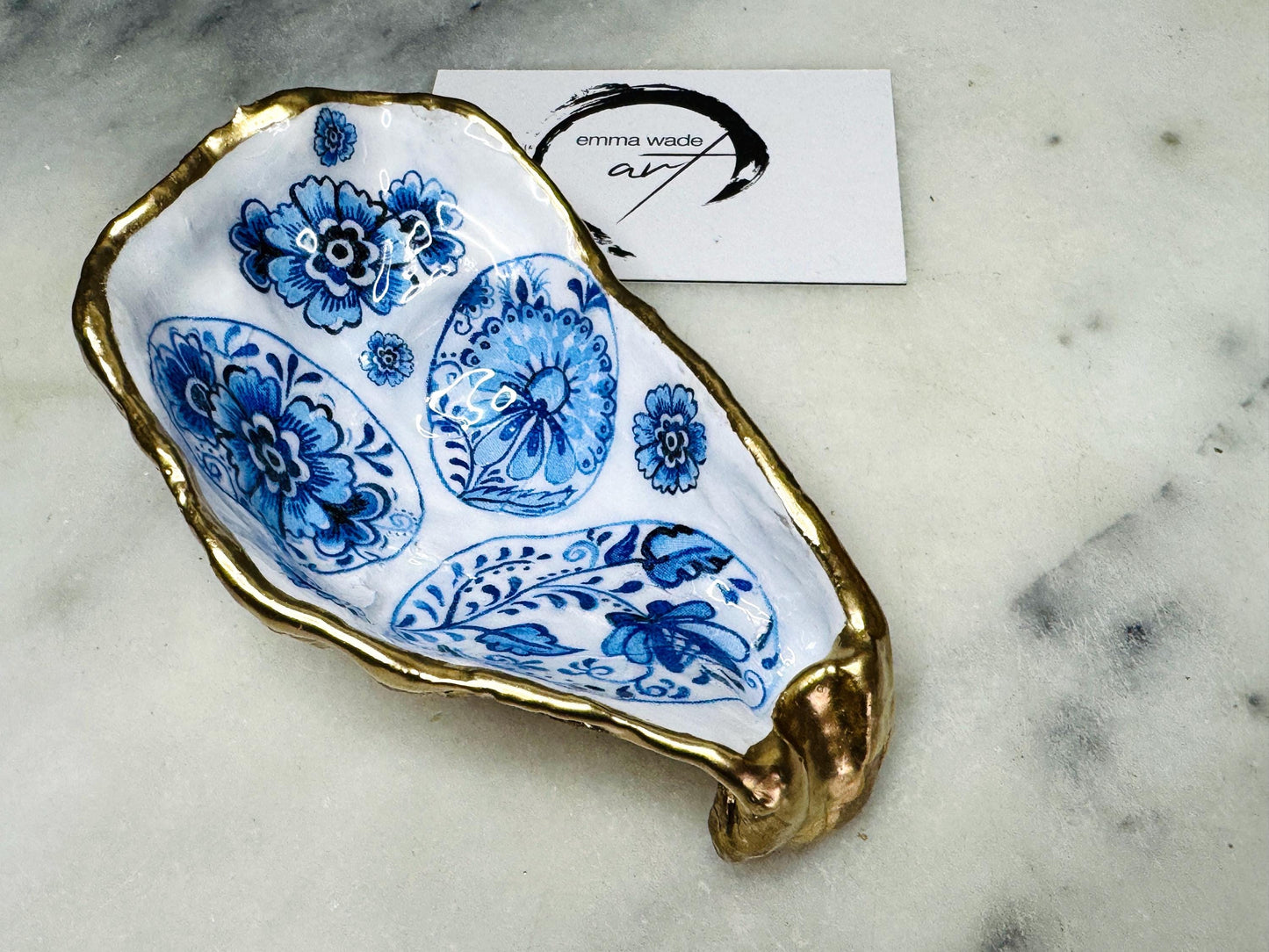 Handmade Dorset oyster shell trinket dish featuring a vintage blue and white decorative egg design with intricate floral porcelain style detailing. Oyster shell is hand gilded with liquid gold leaf edging and sealed in glossy crystal clear resin. Elegant coastal jewelry holder, ring dish, trinket tray, or decorative keepsake inspired by antique ceramics and vintage decor styling. Luxury handmade shell art photographed on a marble background.