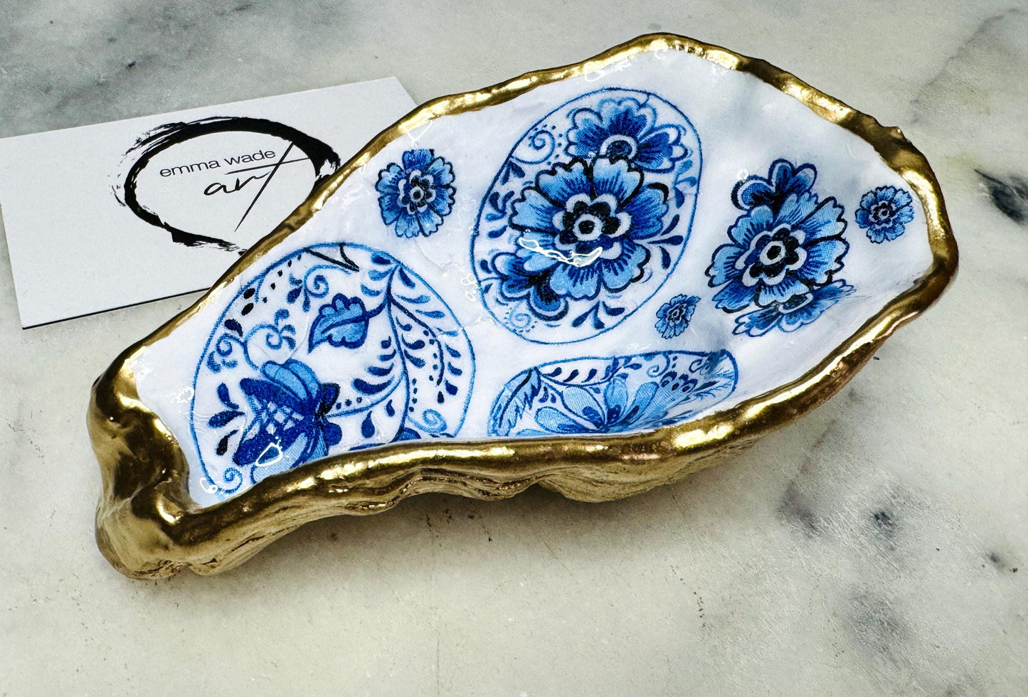 Handmade Dorset oyster shell trinket dish featuring a vintage blue and white decorative egg design with intricate floral porcelain style detailing. Oyster shell is hand gilded with liquid gold leaf edging and sealed in glossy crystal clear resin. Elegant coastal jewelry holder, ring dish, trinket tray, or decorative keepsake inspired by antique ceramics and vintage decor styling. Luxury handmade shell art photographed on a marble background.