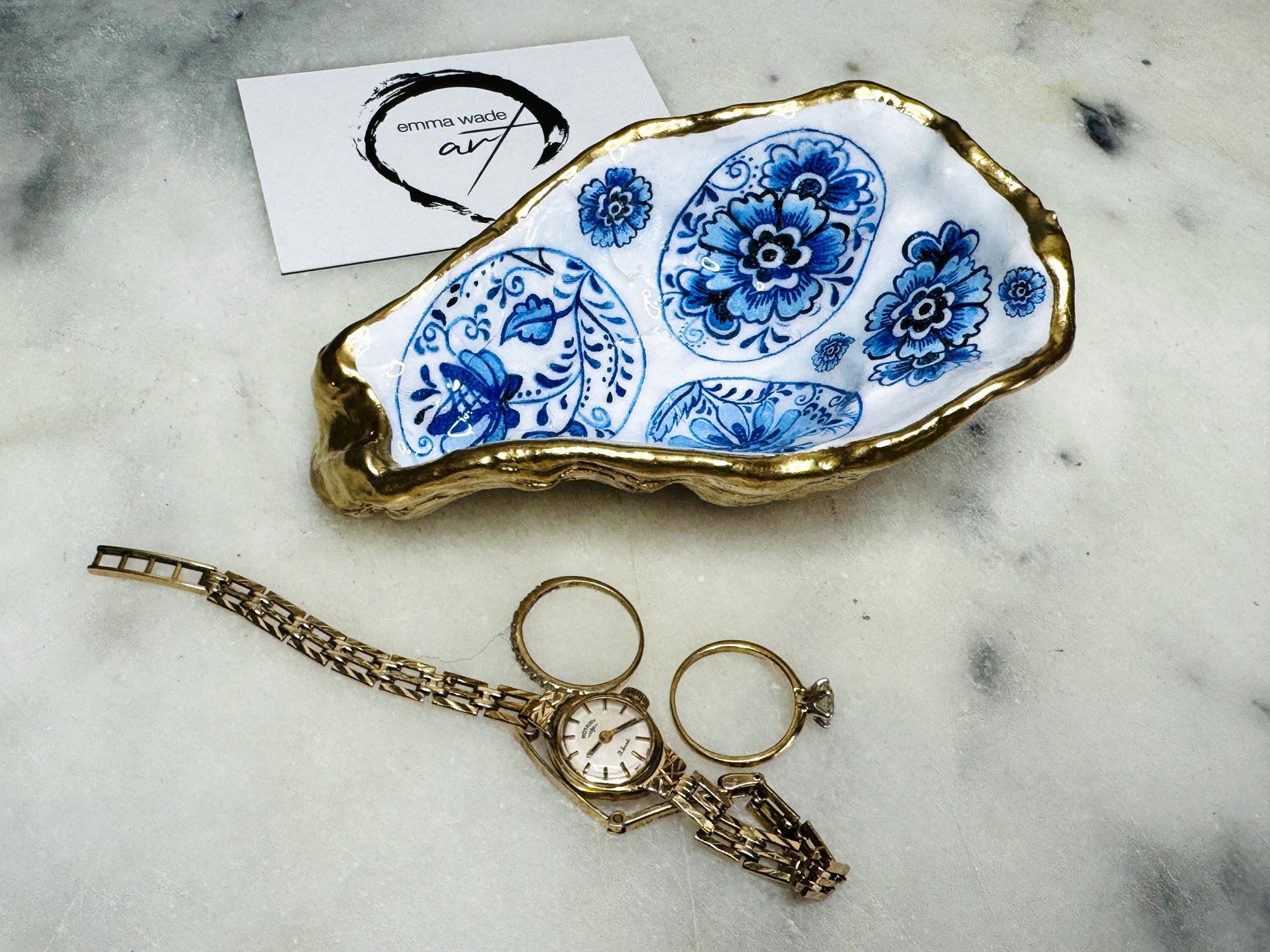 Handmade Dorset oyster shell trinket dish featuring a vintage blue and white decorative egg design with intricate floral porcelain style detailing. Oyster shell is hand gilded with liquid gold leaf edging and sealed in glossy crystal clear resin. Elegant coastal jewelry holder, ring dish, trinket tray, or decorative keepsake inspired by antique ceramics and vintage decor styling. Luxury handmade shell art photographed on a marble background.