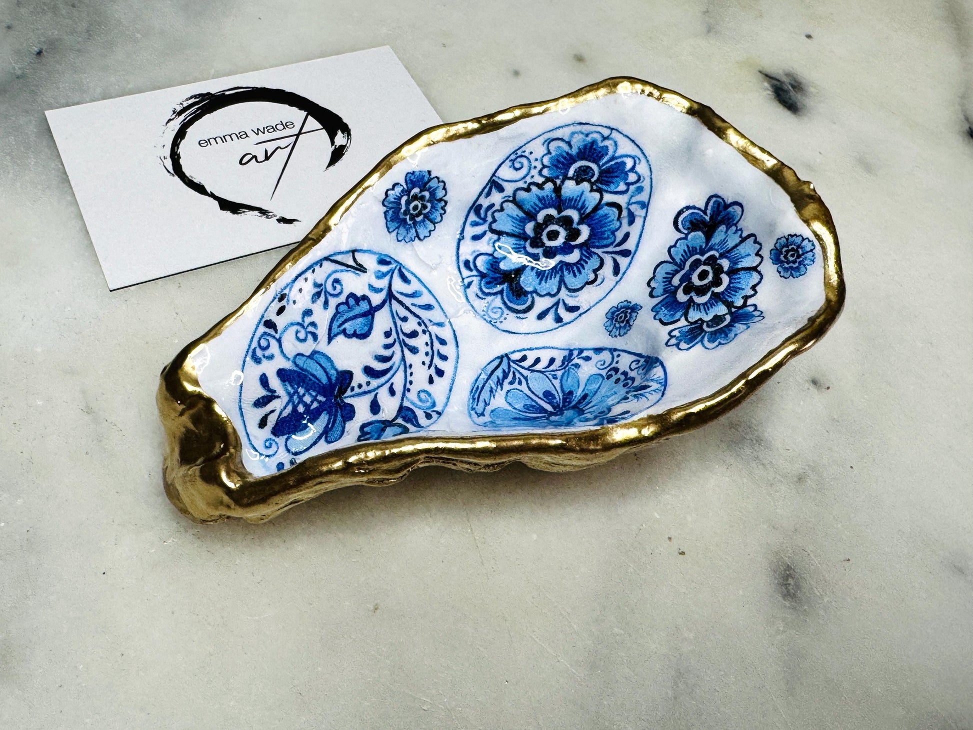 Handmade Dorset oyster shell trinket dish featuring a vintage blue and white decorative egg design with intricate floral porcelain style detailing. Oyster shell is hand gilded with liquid gold leaf edging and sealed in glossy crystal clear resin. Elegant coastal jewelry holder, ring dish, trinket tray, or decorative keepsake inspired by antique ceramics and vintage decor styling. Luxury handmade shell art photographed on a marble background.