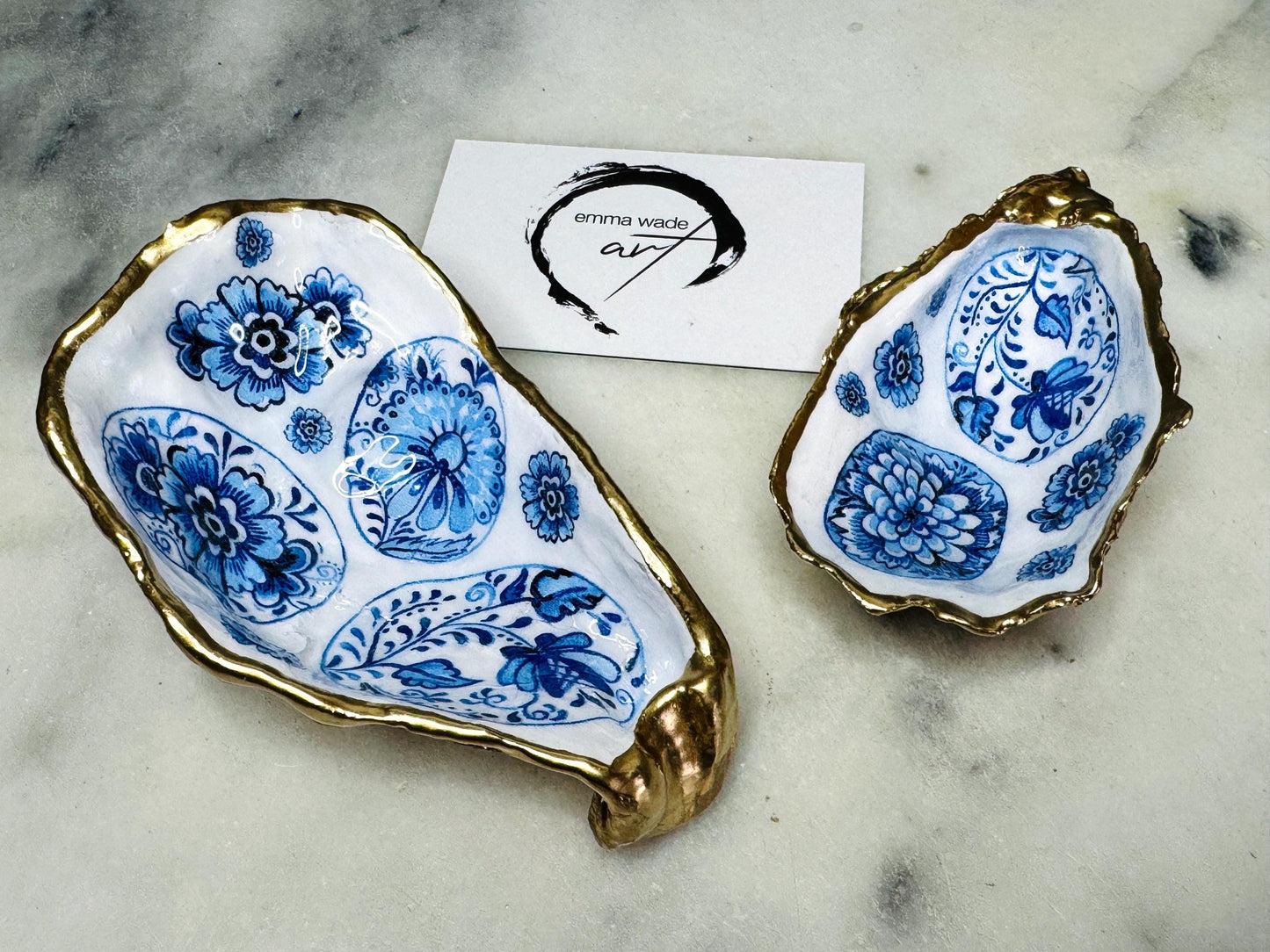 Handmade Dorset oyster shell trinket dish featuring a vintage blue and white decorative egg design with intricate floral porcelain style detailing. Oyster shell is hand gilded with liquid gold leaf edging and sealed in glossy crystal clear resin. Elegant coastal jewelry holder, ring dish, trinket tray, or decorative keepsake inspired by antique ceramics and vintage decor styling. Luxury handmade shell art photographed on a marble background.