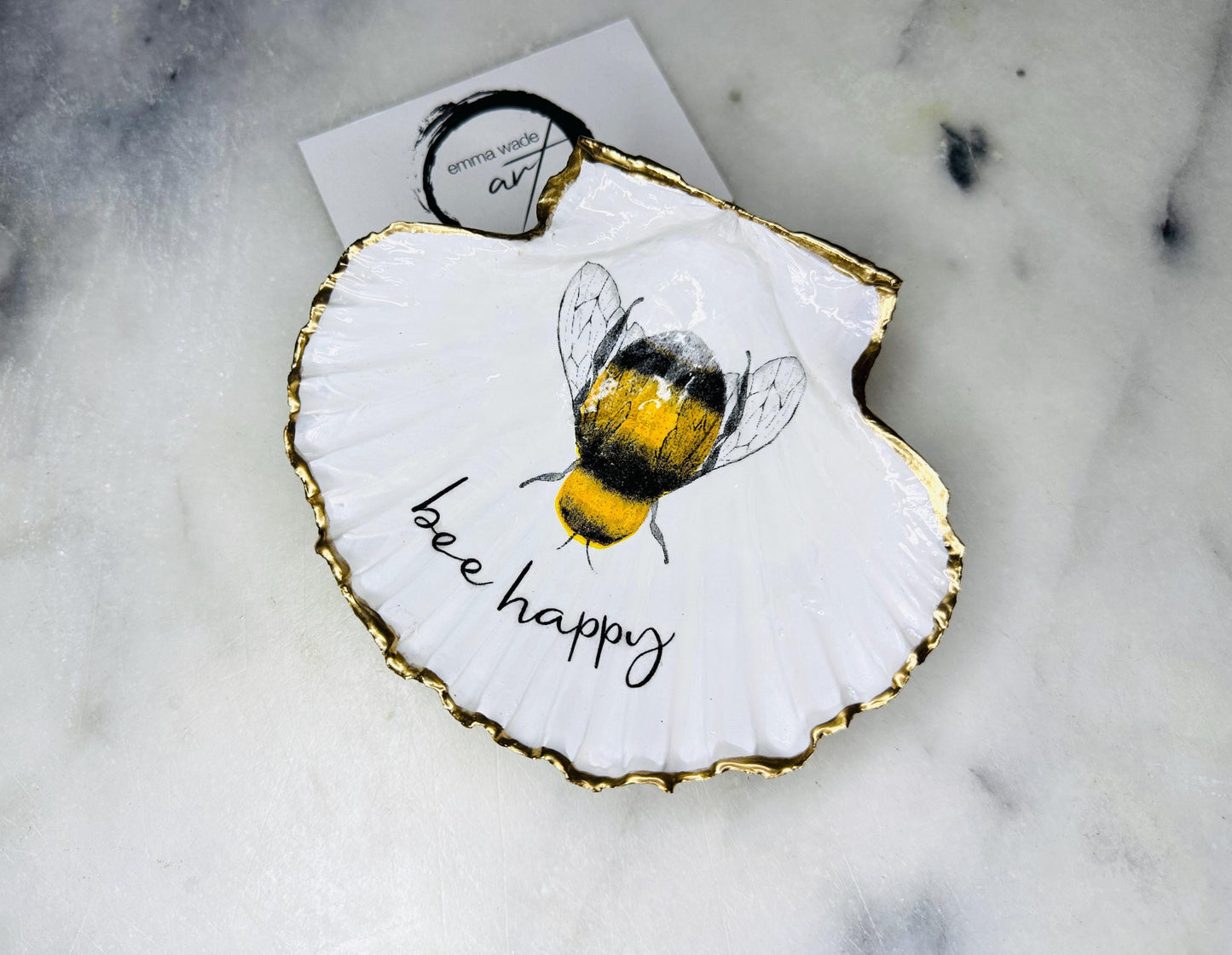 Handmade scallop shell trinket dish featuring a bumblebee illustration with gold leaf edging and glossy resin finish. Coastal jewellery dish in natural white shell with bee artwork, displayed as a ring holder and handmade bee lover gift