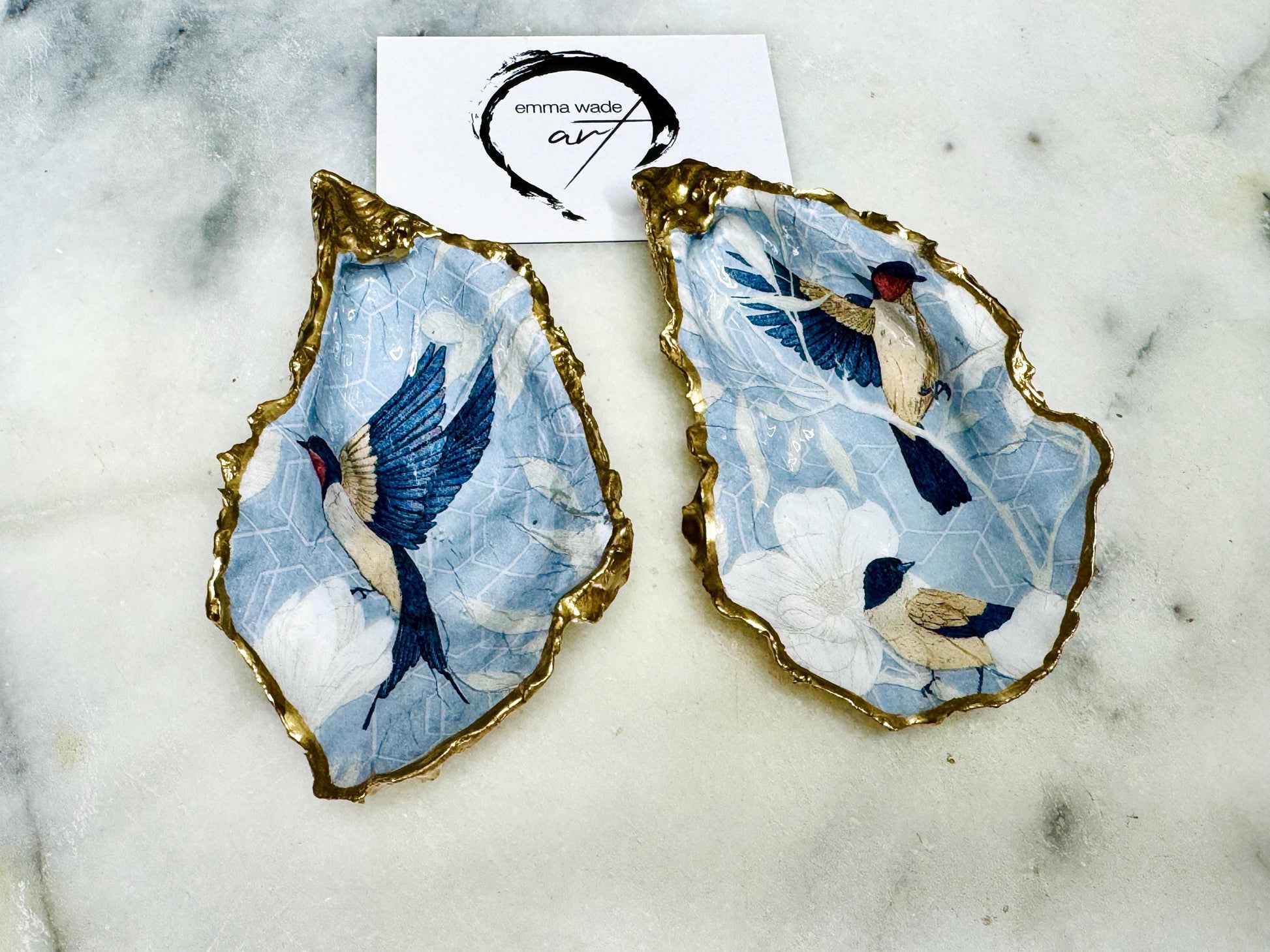 Handmade blue bird oyster shell salt and pepper dishes shown as a matching pair with small gold spoons, featuring a delicate bird design and hand-gilded gold leaf edges. Crafted from natural Dorset oyster shells and sealed with a glossy finish, these coastal-inspired pinch pots are perfect for table decor, weddings, or a unique hostess gift. Each shell is one of a kind and made by hand.