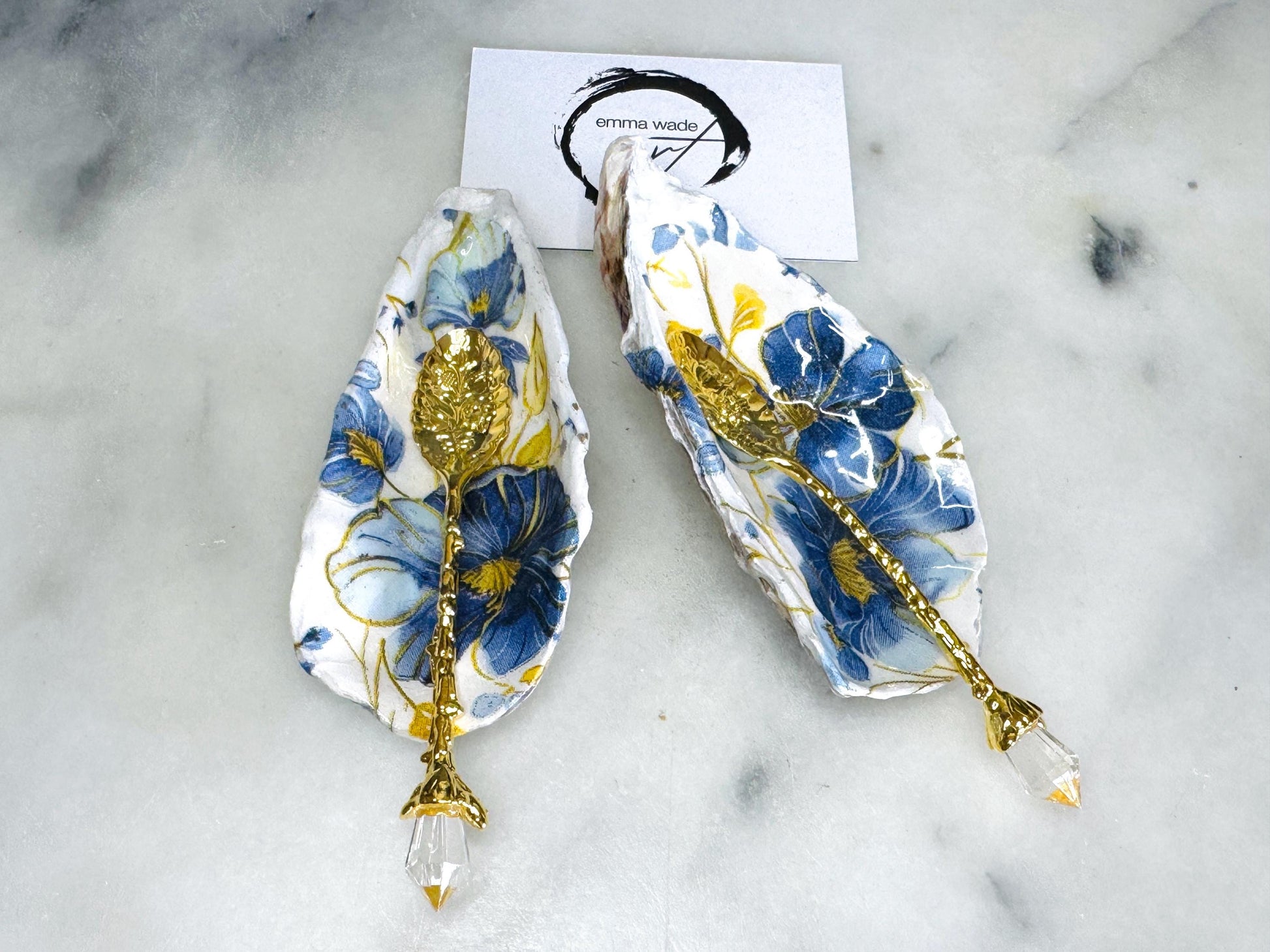 Handmade blue and yellow floral oyster shell salt and pepper dishes shown as a matching pair, crafted from natural Dorset shells and styled with small gold spoons. Coastal-inspired pinch pots featuring a decorative floral design with natural shell backs, perfect for table decor, weddings, or a unique hostess gift. Each shell is one of a kind, finished by hand, and ideal for adding colour and elegance to a dining table.
