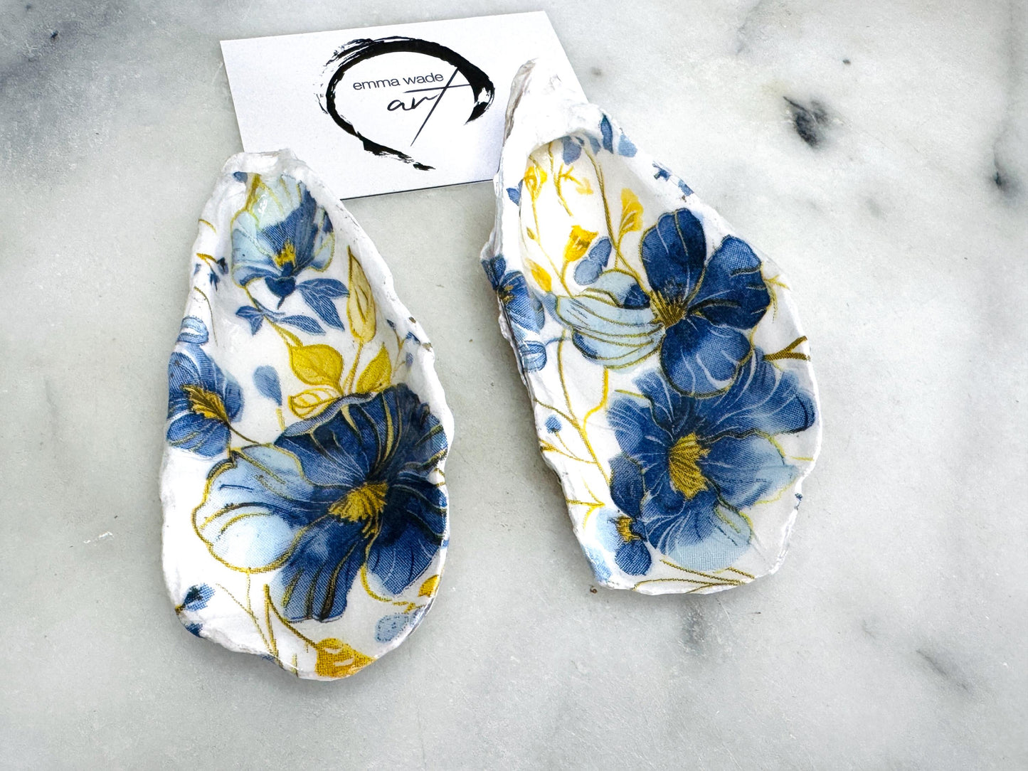 Handmade blue and yellow floral oyster shell salt and pepper dishes shown as a matching pair, crafted from natural Dorset shells and styled with small gold spoons. Coastal-inspired pinch pots featuring a decorative floral design with natural shell backs, perfect for table decor, weddings, or a unique hostess gift. Each shell is one of a kind, finished by hand, and ideal for adding colour and elegance to a dining table.