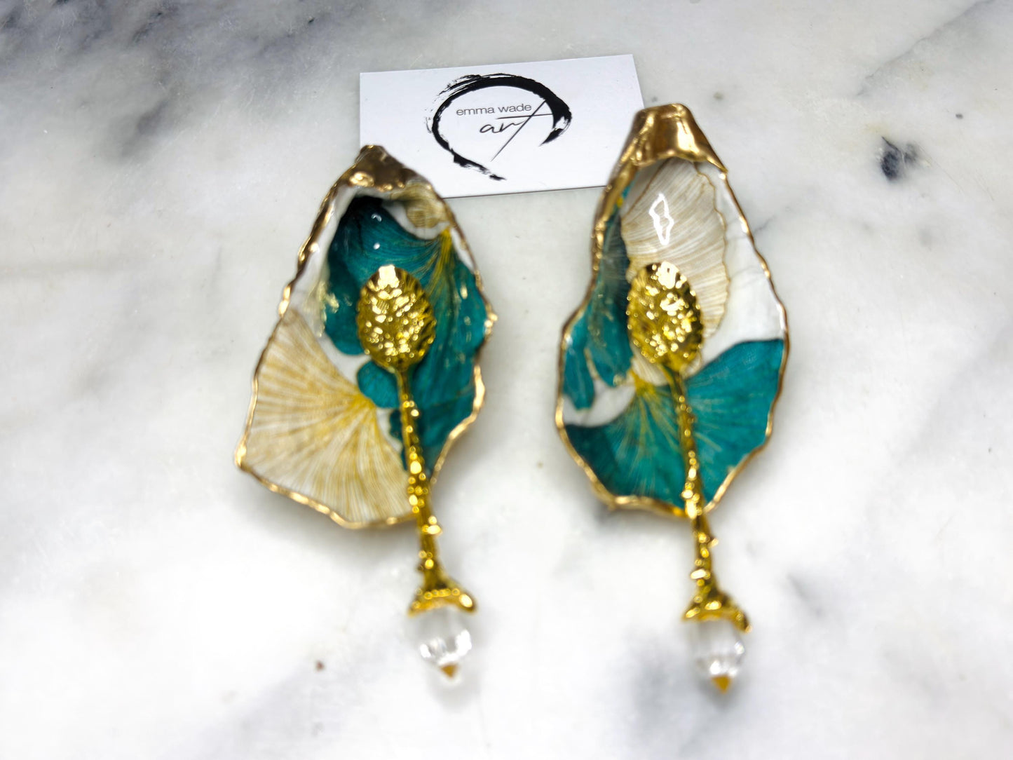 A pair of green and gold Japanese Ginkgo oyster shell salt and pepper pinch pots with floral botanical leaf design, hand gilded with liquid gold leaf and sealed in glossy resin. Coastal Dorset oyster shell dishes styled with matching gold crystal spoons on a marble background. Handmade luxury coastal dining décor and wedding gift.