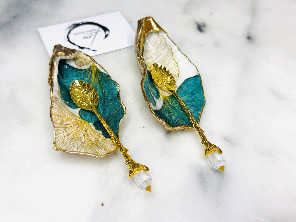 A pair of green and gold Japanese Ginkgo oyster shell salt and pepper pinch pots with floral botanical leaf design, hand gilded with liquid gold leaf and sealed in glossy resin. Coastal Dorset oyster shell dishes styled with matching gold crystal spoons on a marble background. Handmade luxury coastal dining décor and wedding gift.