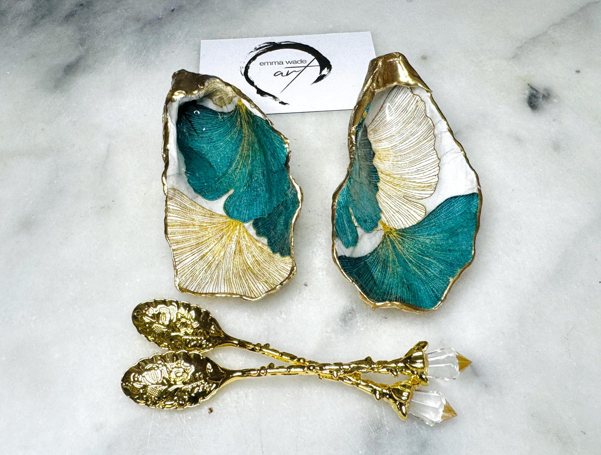 A pair of green and gold Japanese Ginkgo oyster shell salt and pepper pinch pots with floral botanical leaf design, hand gilded with liquid gold leaf and sealed in glossy resin. Coastal Dorset oyster shell dishes styled with matching gold crystal spoons on a marble background. Handmade luxury coastal dining décor and wedding gift.