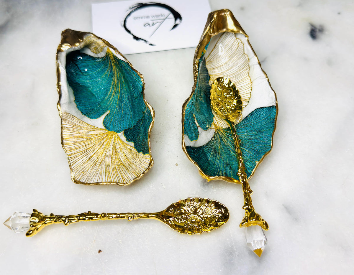 A pair of green and gold Japanese Ginkgo oyster shell salt and pepper pinch pots with floral botanical leaf design, hand gilded with liquid gold leaf and sealed in glossy resin. Coastal Dorset oyster shell dishes styled with matching gold crystal spoons on a marble background. Handmade luxury coastal dining décor and wedding gift.