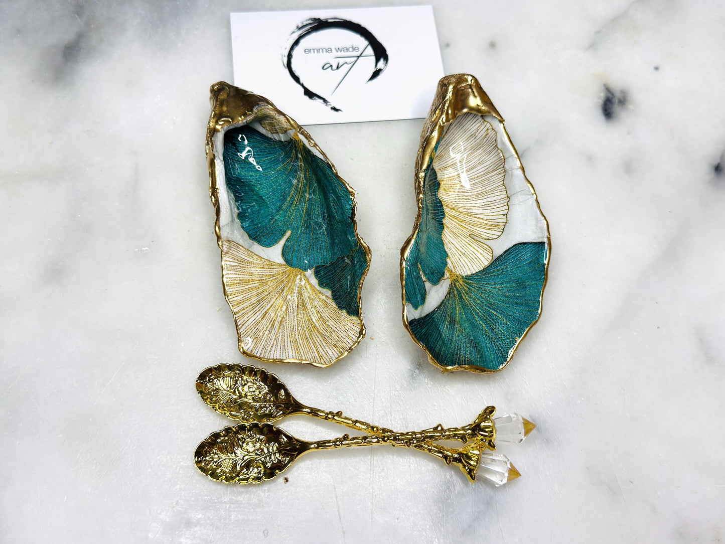 A pair of green and gold Japanese Ginkgo oyster shell salt and pepper pinch pots with floral botanical leaf design, hand gilded with liquid gold leaf and sealed in glossy resin. Coastal Dorset oyster shell dishes styled with matching gold crystal spoons on a marble background. Handmade luxury coastal dining décor and wedding gift.