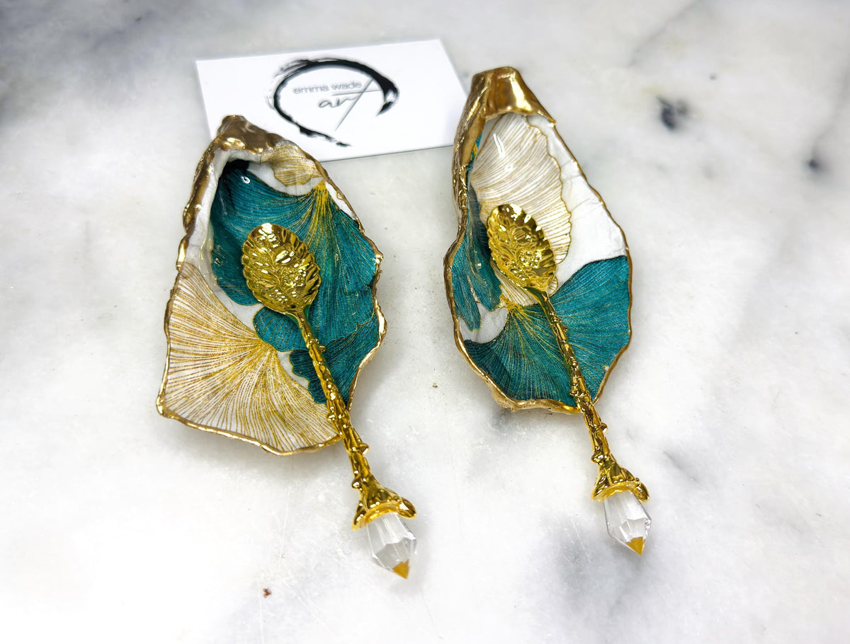 A pair of green and gold Japanese Ginkgo oyster shell salt and pepper pinch pots with floral botanical leaf design, hand gilded with liquid gold leaf and sealed in glossy resin. Coastal Dorset oyster shell dishes styled with matching gold crystal spoons on a marble background. Handmade luxury coastal dining décor and wedding gift.