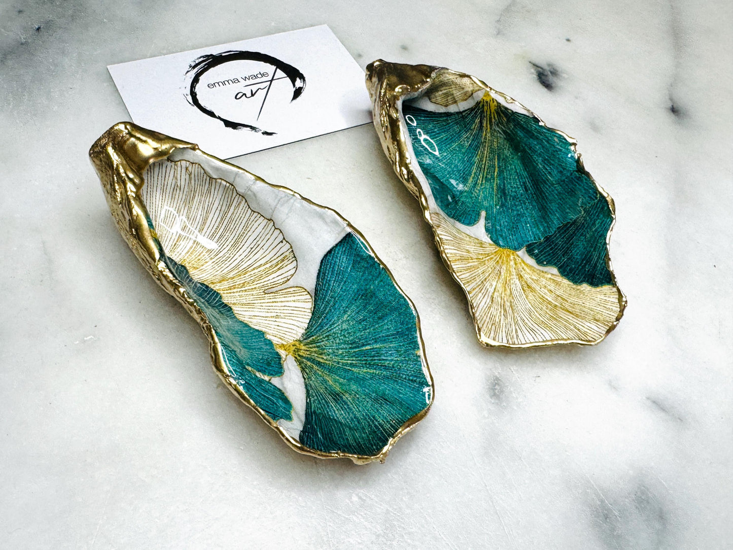 A pair of green and gold Japanese Ginkgo oyster shell salt and pepper pinch pots with floral botanical leaf design, hand gilded with liquid gold leaf and sealed in glossy resin. Coastal Dorset oyster shell dishes styled with matching gold crystal spoons on a marble background. Handmade luxury coastal dining décor and wedding gift.