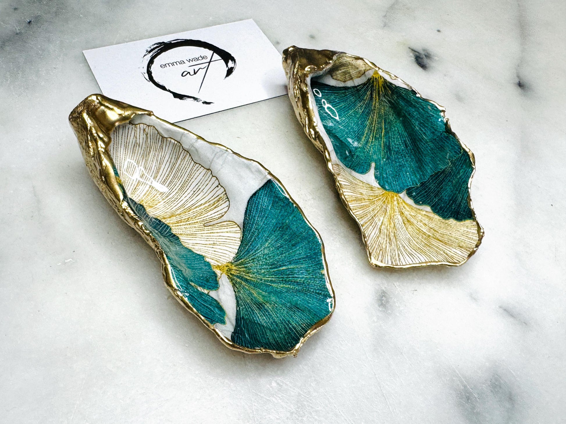 A pair of green and gold Japanese Ginkgo oyster shell salt and pepper pinch pots with floral botanical leaf design, hand gilded with liquid gold leaf and sealed in glossy resin. Coastal Dorset oyster shell dishes styled with matching gold crystal spoons on a marble background. Handmade luxury coastal dining décor and wedding gift.
