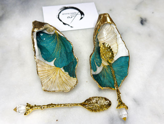 A pair of green and gold Japanese Ginkgo oyster shell salt and pepper pinch pots with floral botanical leaf design, hand gilded with liquid gold leaf and sealed in glossy resin. Coastal Dorset oyster shell dishes styled with matching gold crystal spoons on a marble background. Handmade luxury coastal dining décor and wedding gift.