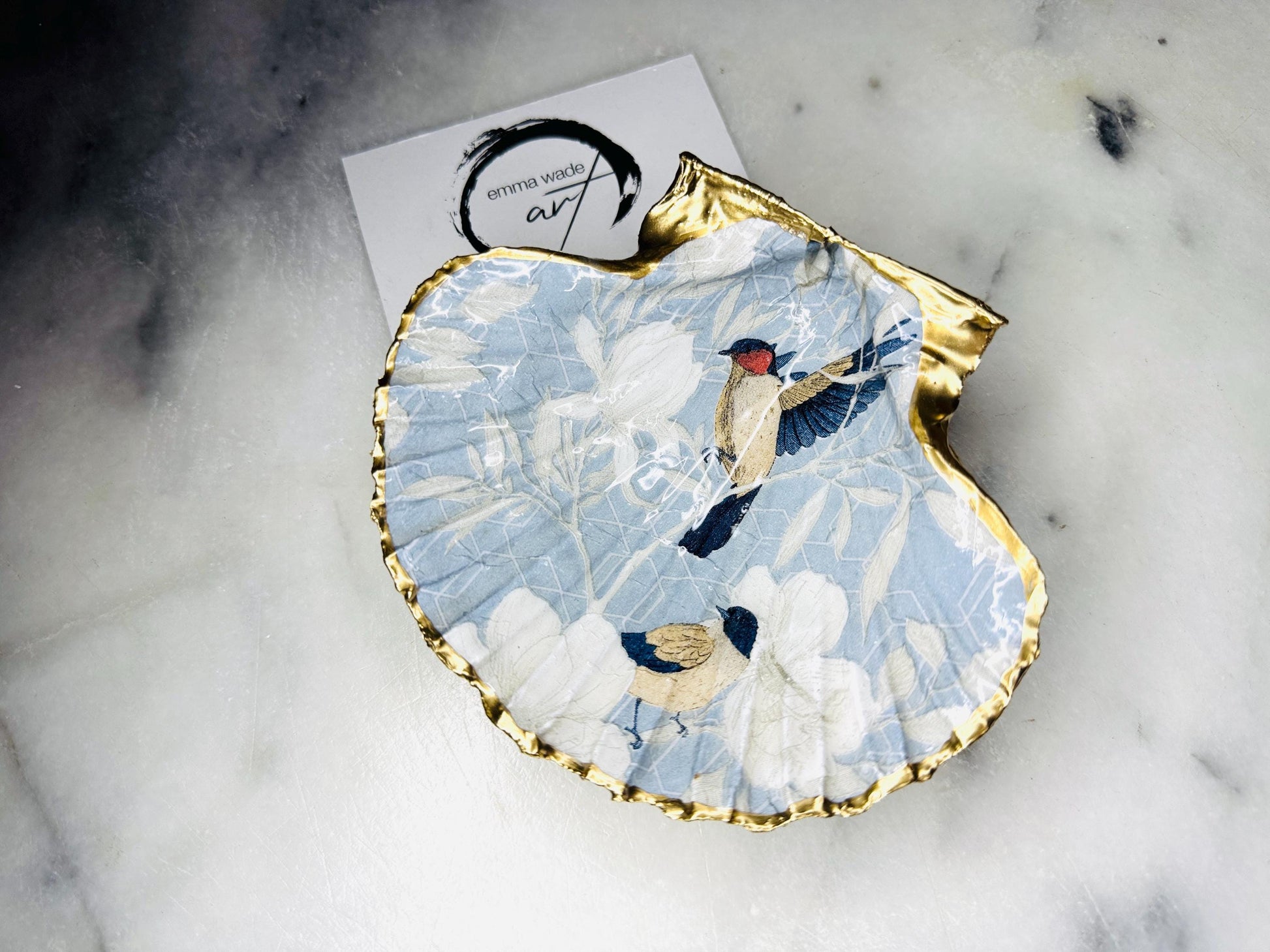 Handmade scallop shell trinket dish featuring a Blue Birds of Paradise decoupage design, finished with real gold leaf and sealed with glossy resin. Crafted from a natural Dorset scallop shell and shown as a jewellery and ring holder on a light background. Coastal-inspired home decor, perfect for holding rings, earrings and small keepsakes, with each shell one of a kind and finished by hand.