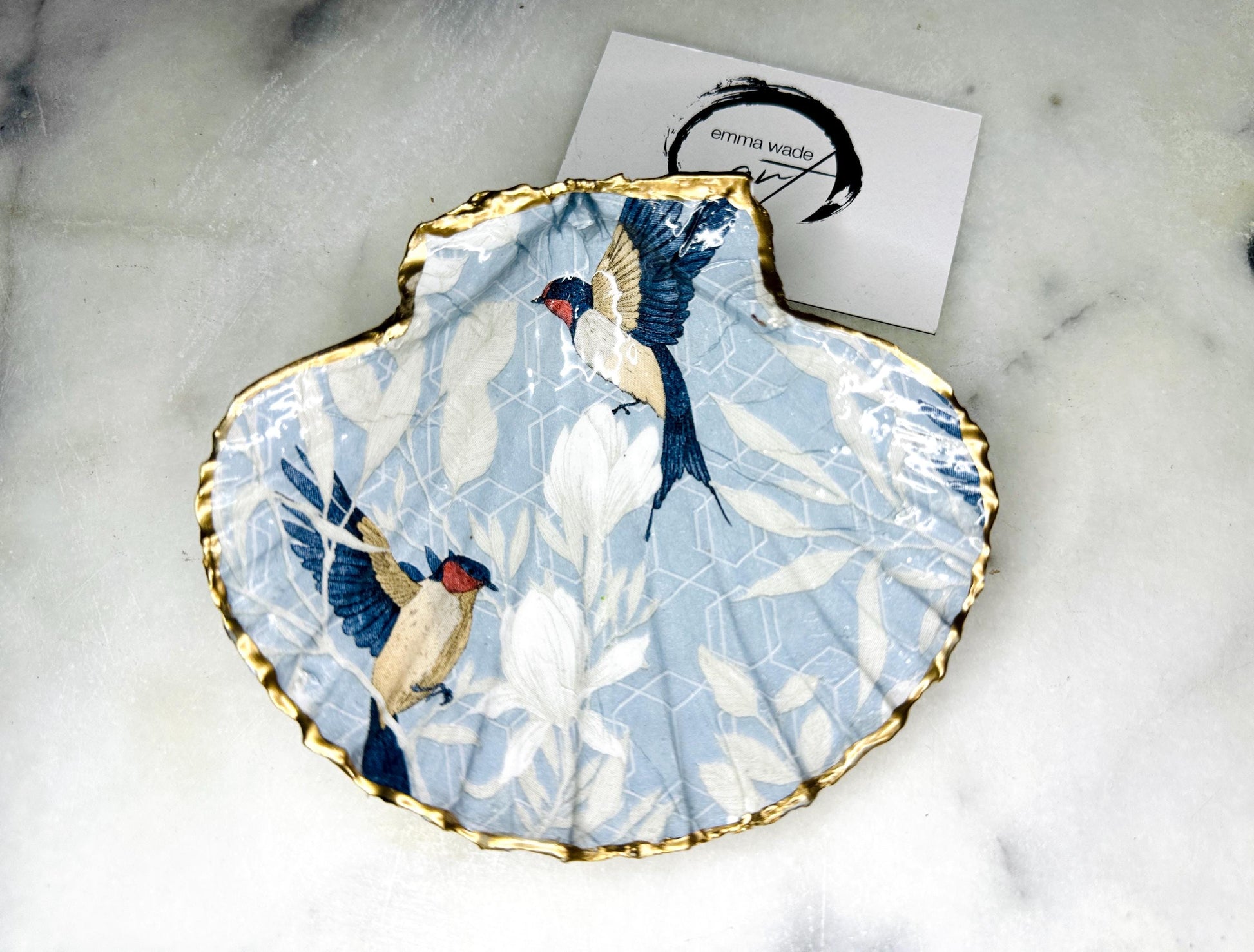 Handmade scallop shell trinket dish featuring a Blue Birds of Paradise decoupage design, finished with real gold leaf and sealed with glossy resin. Crafted from a natural Dorset scallop shell and shown as a jewellery and ring holder on a light background. Coastal-inspired home decor, perfect for holding rings, earrings and small keepsakes, with each shell one of a kind and finished by hand.