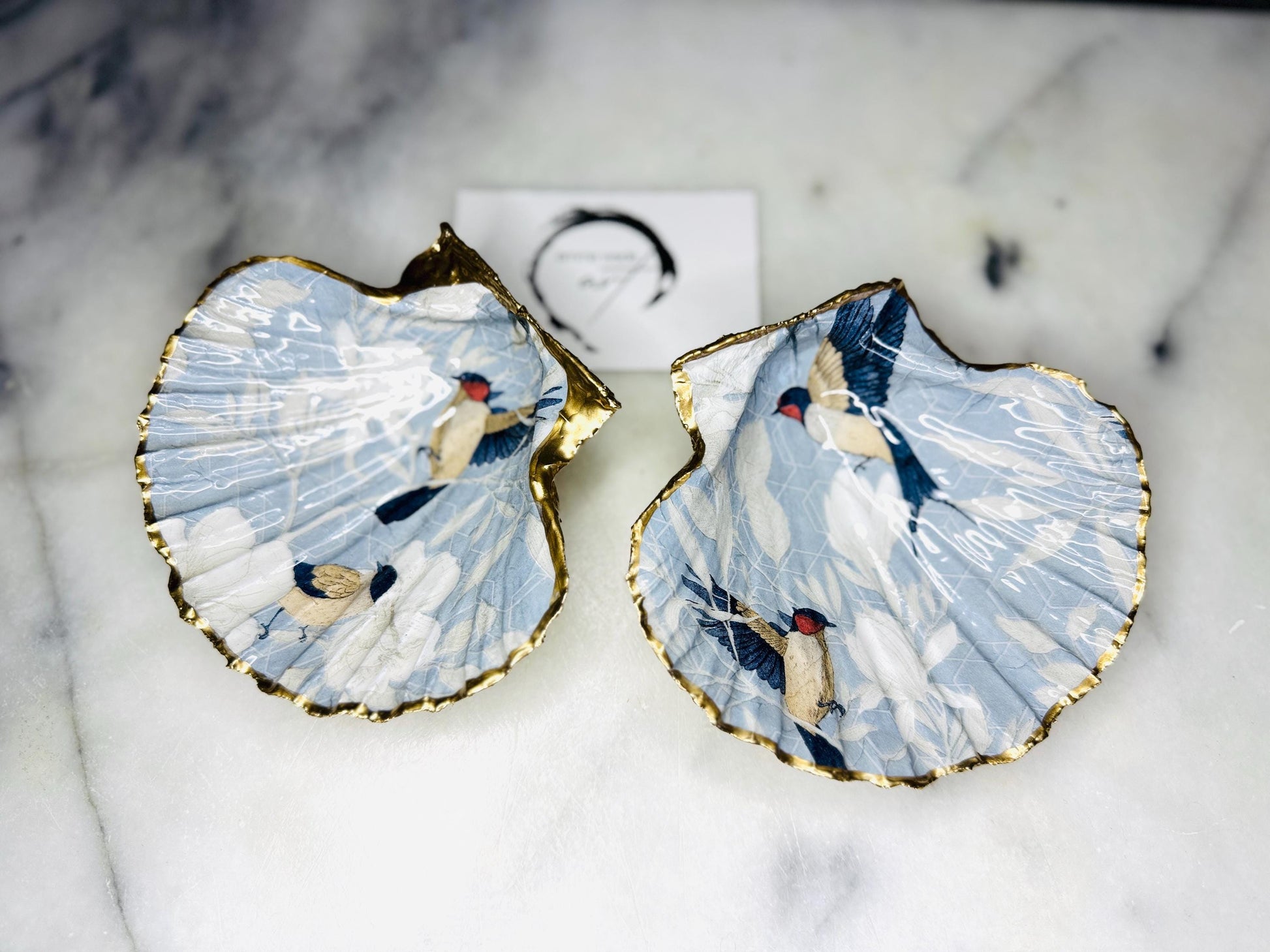 Handmade scallop shell trinket dish featuring a Blue Birds of Paradise decoupage design, finished with real gold leaf and sealed with glossy resin. Crafted from a natural Dorset scallop shell and shown as a jewellery and ring holder on a light background. Coastal-inspired home decor, perfect for holding rings, earrings and small keepsakes, with each shell one of a kind and finished by hand.