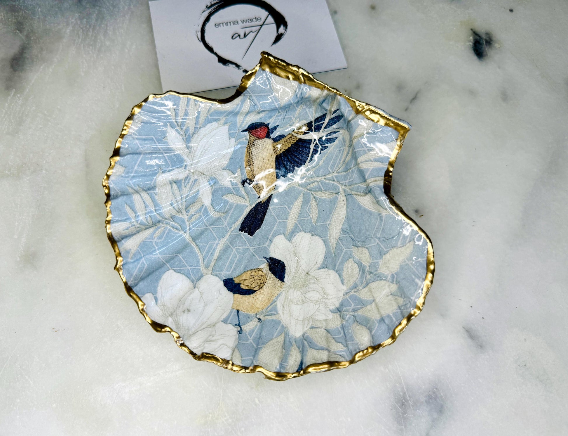 Handmade scallop shell trinket dish featuring a Blue Birds of Paradise decoupage design, finished with real gold leaf and sealed with glossy resin. Crafted from a natural Dorset scallop shell and shown as a jewellery and ring holder on a light background. Coastal-inspired home decor, perfect for holding rings, earrings and small keepsakes, with each shell one of a kind and finished by hand.
