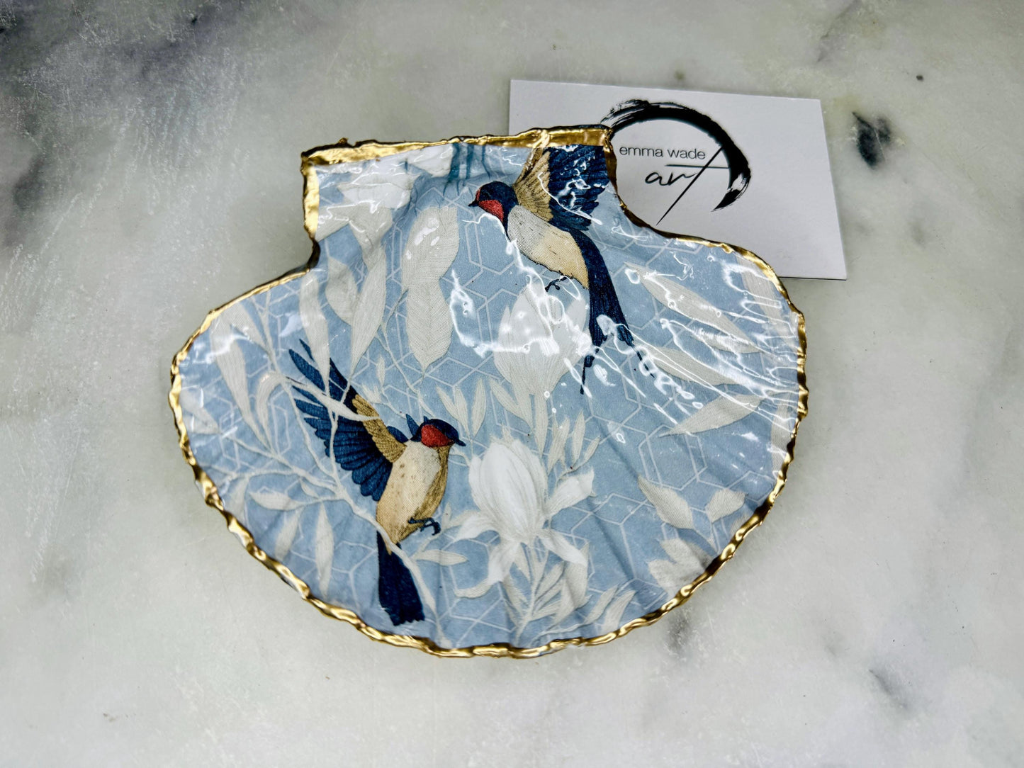 Handmade scallop shell trinket dish featuring a Blue Birds of Paradise decoupage design, finished with real gold leaf and sealed with glossy resin. Crafted from a natural Dorset scallop shell and shown as a jewellery and ring holder on a light background. Coastal-inspired home decor, perfect for holding rings, earrings and small keepsakes, with each shell one of a kind and finished by hand.