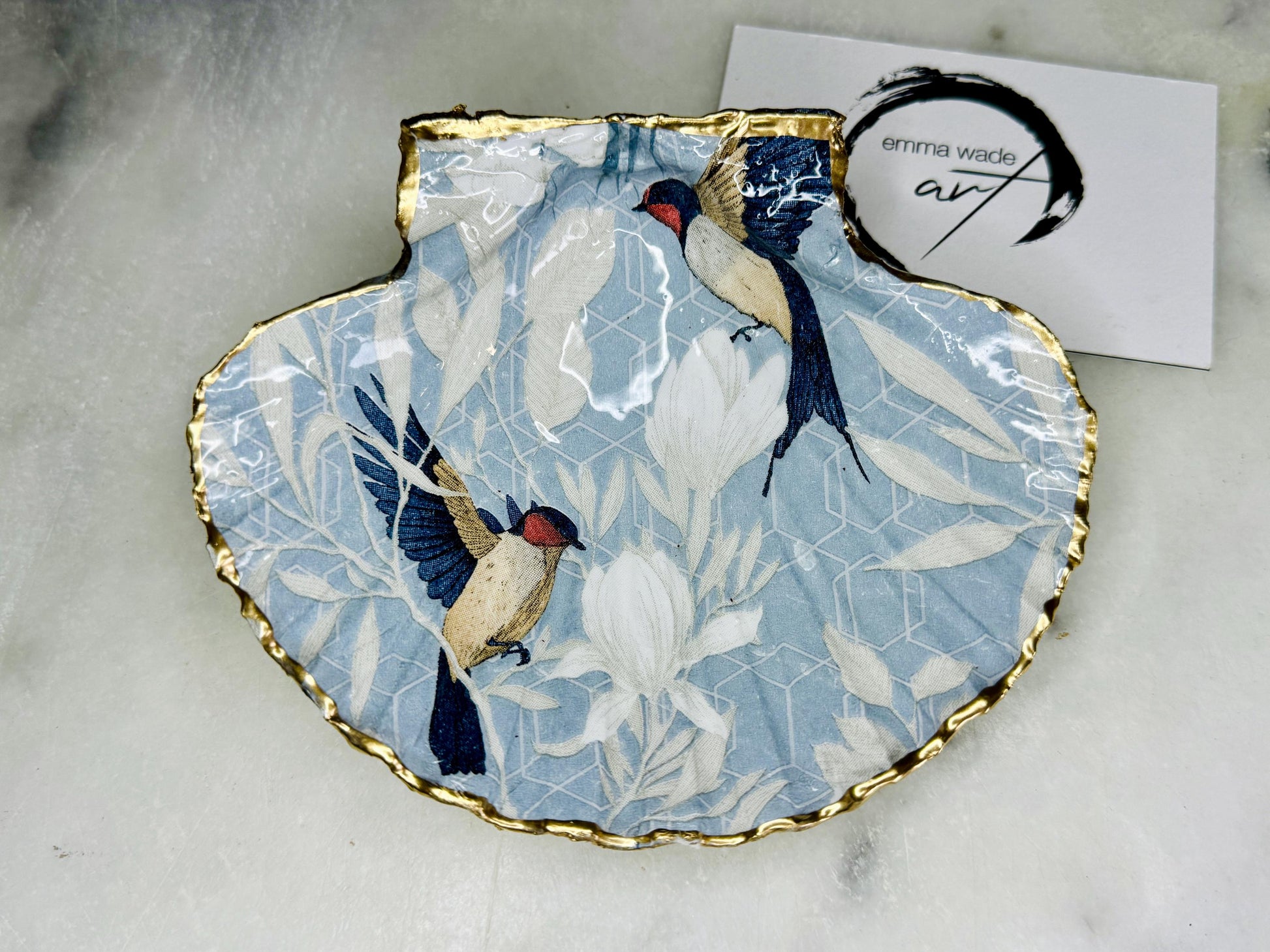 Handmade scallop shell trinket dish featuring a Blue Birds of Paradise decoupage design, finished with real gold leaf and sealed with glossy resin. Crafted from a natural Dorset scallop shell and shown as a jewellery and ring holder on a light background. Coastal-inspired home decor, perfect for holding rings, earrings and small keepsakes, with each shell one of a kind and finished by hand.
