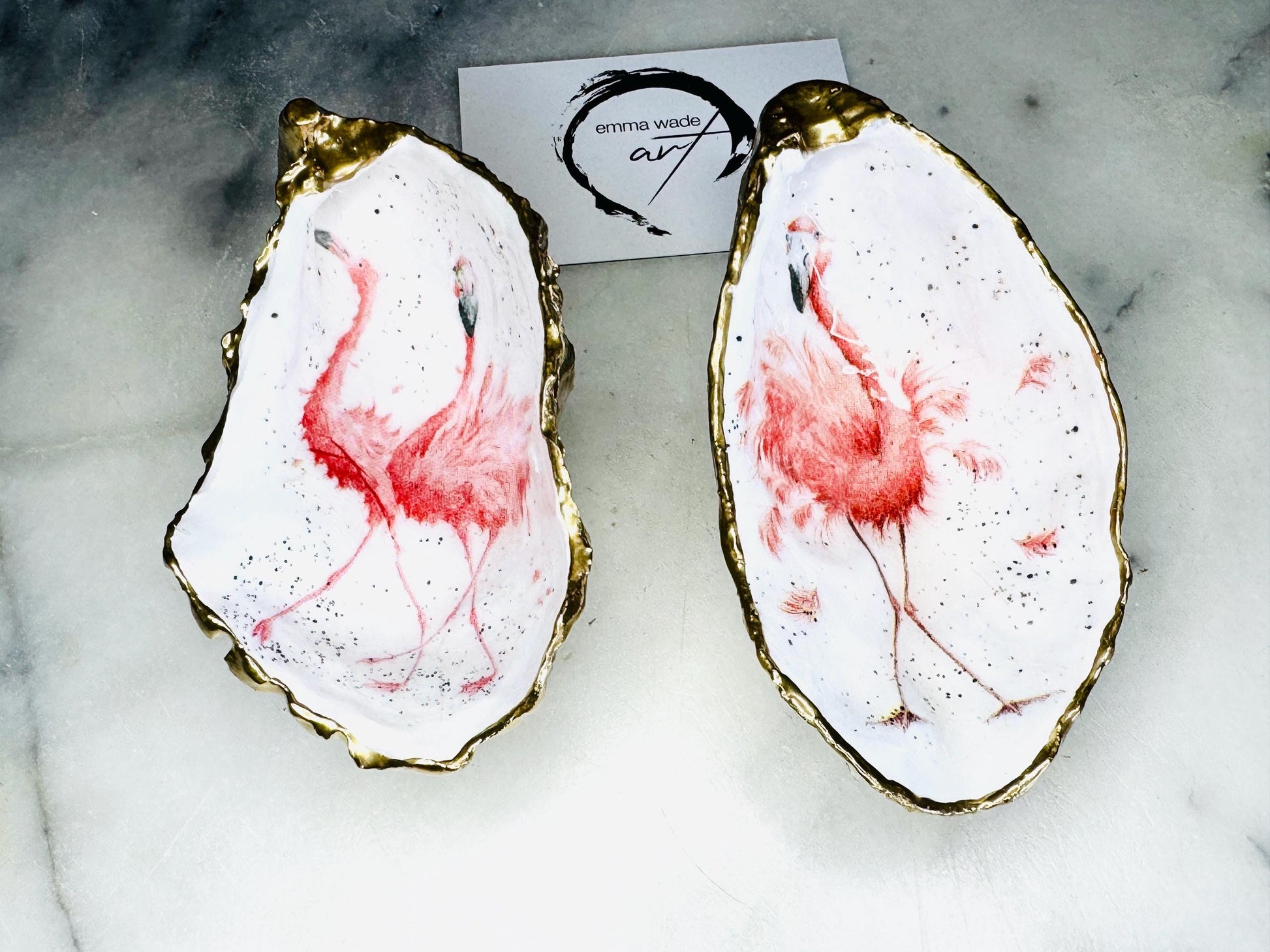 Handmade Dorset oyster shell trinket dishes featuring elegant pink flamingo bird artwork with hand-painted gold leaf edging and glossy crystal clear resin. Coastal flamingo jewellery holders styled on a white marble background with tropical blush pink tones and luxury coastal home décor styling.