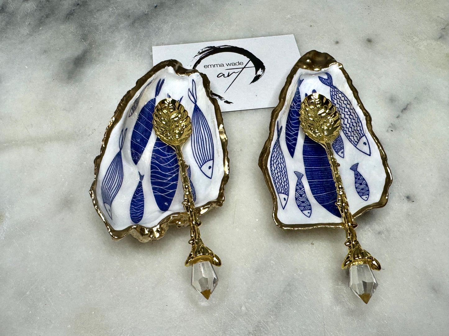 Pair of handmade Dorset oyster shell salt and pepper pinch pots decorated with elegant blue and white fish artwork, finished with hand applied liquid gold leaf edging and sealed in glossy food-safe resin. Coastal shell cruet dishes styled with optional gold crystal spoons on a marble background. Luxury nautical kitchen decor inspired by the sea, perfect for beach house dining, dinner parties, weddings, anniversaries, and coastal home gifts.