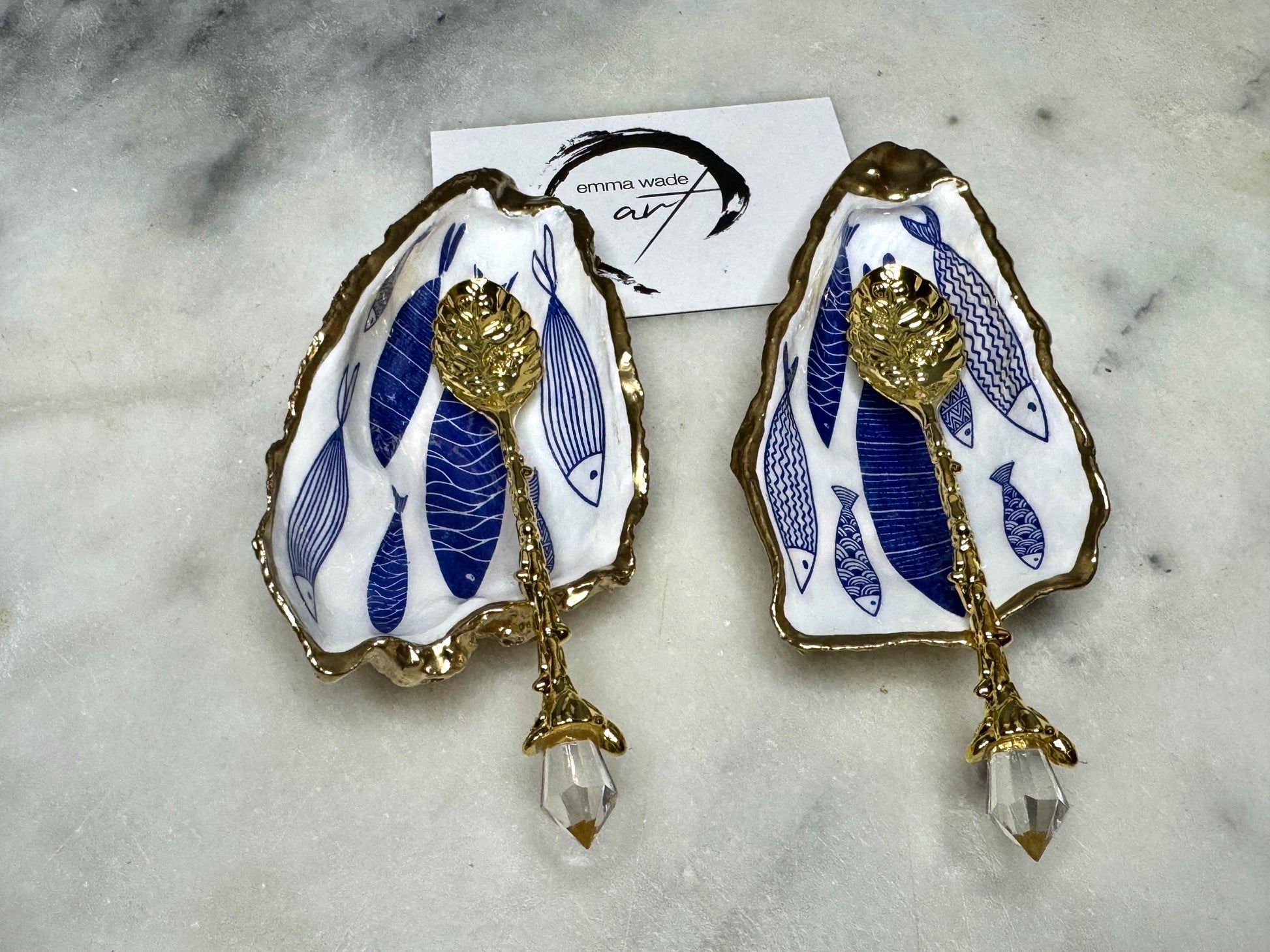 Pair of handmade Dorset oyster shell salt and pepper pinch pots decorated with elegant blue and white fish artwork, finished with hand applied liquid gold leaf edging and sealed in glossy food-safe resin. Coastal shell cruet dishes styled with optional gold crystal spoons on a marble background. Luxury nautical kitchen decor inspired by the sea, perfect for beach house dining, dinner parties, weddings, anniversaries, and coastal home gifts.