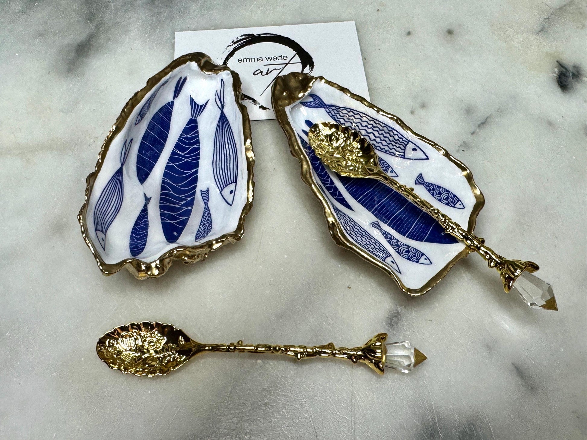 Pair of handmade Dorset oyster shell salt and pepper pinch pots decorated with elegant blue and white fish artwork, finished with hand applied liquid gold leaf edging and sealed in glossy food-safe resin. Coastal shell cruet dishes styled with optional gold crystal spoons on a marble background. Luxury nautical kitchen decor inspired by the sea, perfect for beach house dining, dinner parties, weddings, anniversaries, and coastal home gifts.