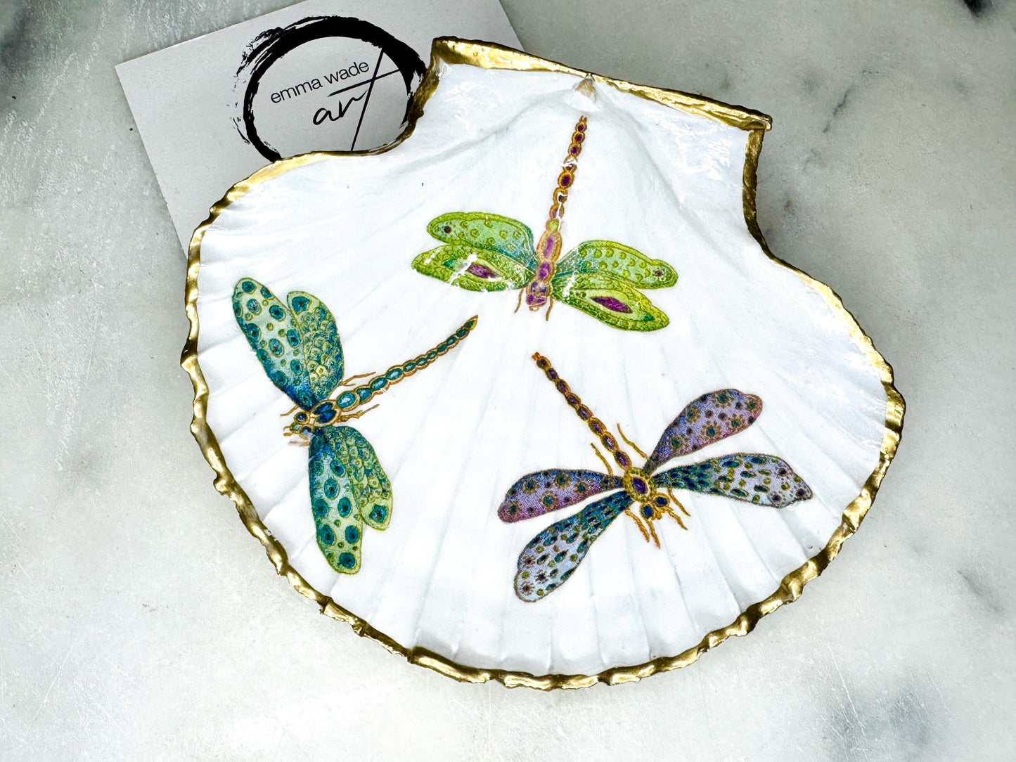 Handmade dragonfly scallop shell trinket dish with gold leaf edging and glossy resin finish. Coastal jewellery and ring dish crafted from a natural Dorset scallop shell, featuring colourful dragonfly artwork. Decorative shell bowl photographed on a light background, showing the natural shell shape and shimmering gilded edges, ideal for holding rings, earrings, trinkets or small treasures.