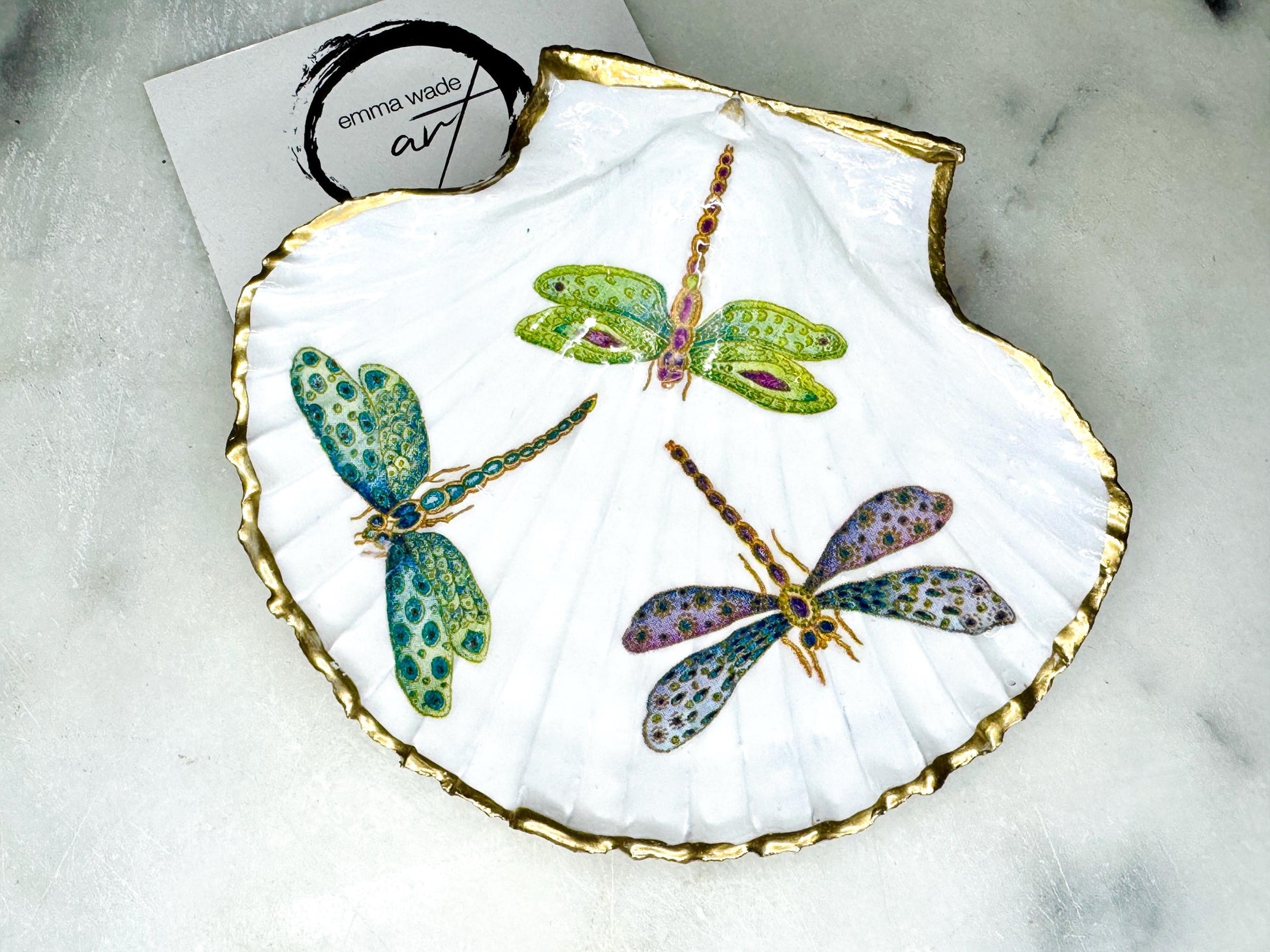 Handmade dragonfly scallop shell trinket dish with gold leaf edging and glossy resin finish. Coastal jewellery and ring dish crafted from a natural Dorset scallop shell, featuring colourful dragonfly artwork. Decorative shell bowl photographed on a light background, showing the natural shell shape and shimmering gilded edges, ideal for holding rings, earrings, trinkets or small treasures.