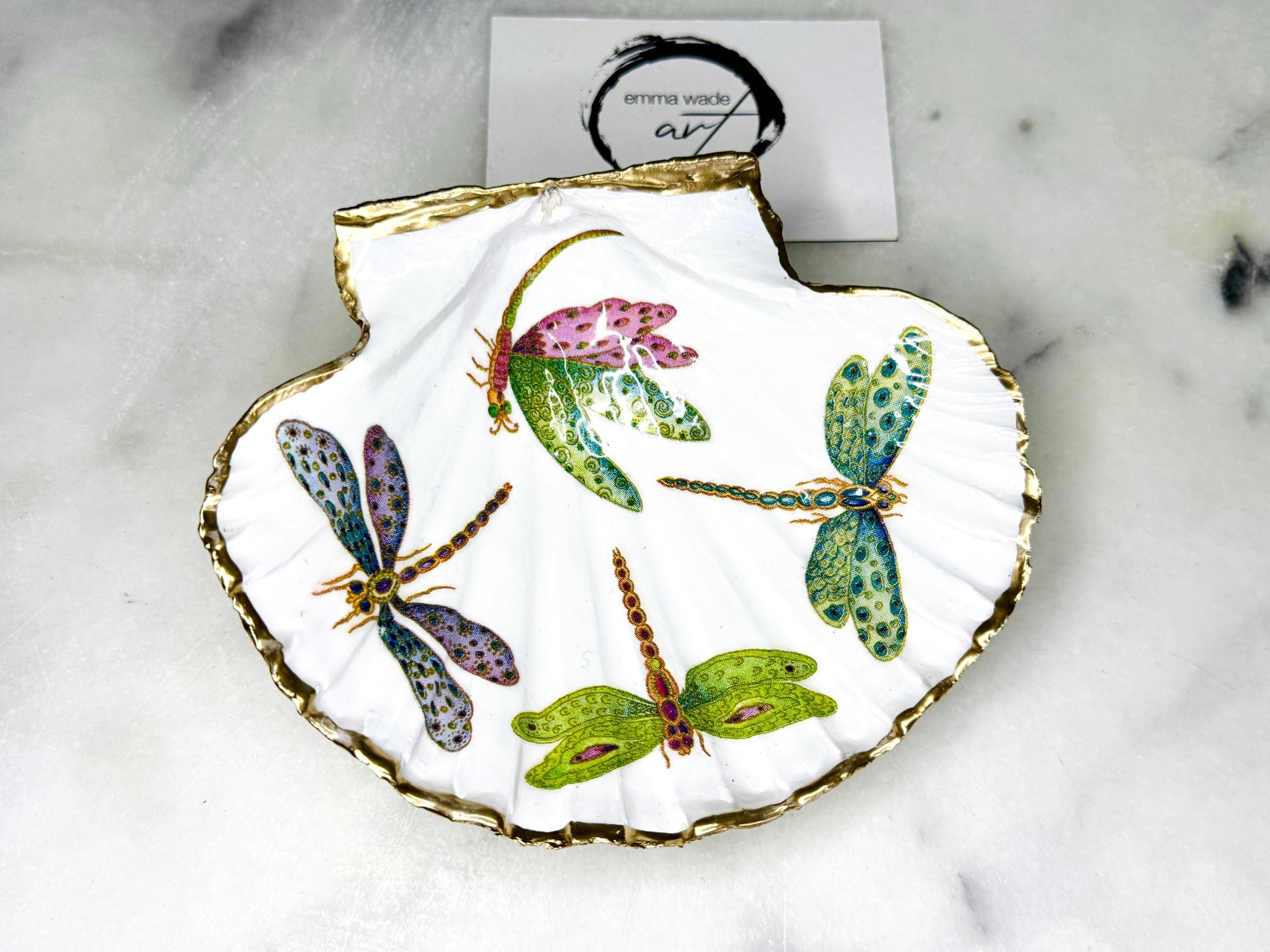 Handmade dragonfly scallop shell trinket dish with gold leaf edging and glossy resin finish. Coastal jewellery and ring dish crafted from a natural Dorset scallop shell, featuring colourful dragonfly artwork. Decorative shell bowl photographed on a light background, showing the natural shell shape and shimmering gilded edges, ideal for holding rings, earrings, trinkets or small treasures.