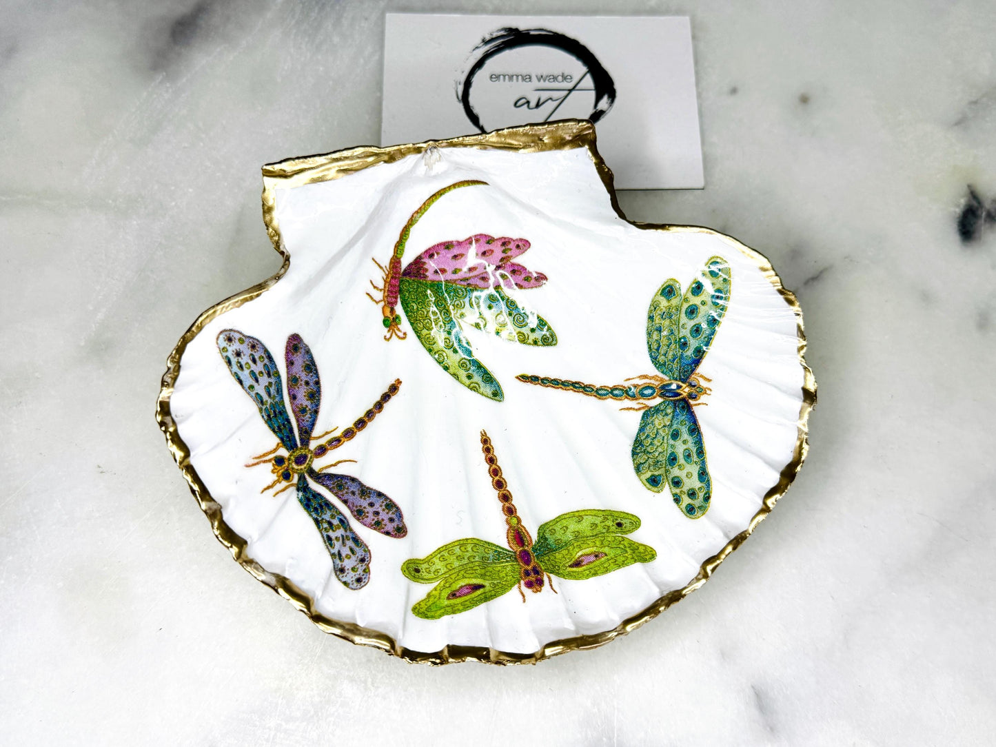 Handmade dragonfly scallop shell trinket dish with gold leaf edging and glossy resin finish. Coastal jewellery and ring dish crafted from a natural Dorset scallop shell, featuring colourful dragonfly artwork. Decorative shell bowl photographed on a light background, showing the natural shell shape and shimmering gilded edges, ideal for holding rings, earrings, trinkets or small treasures.