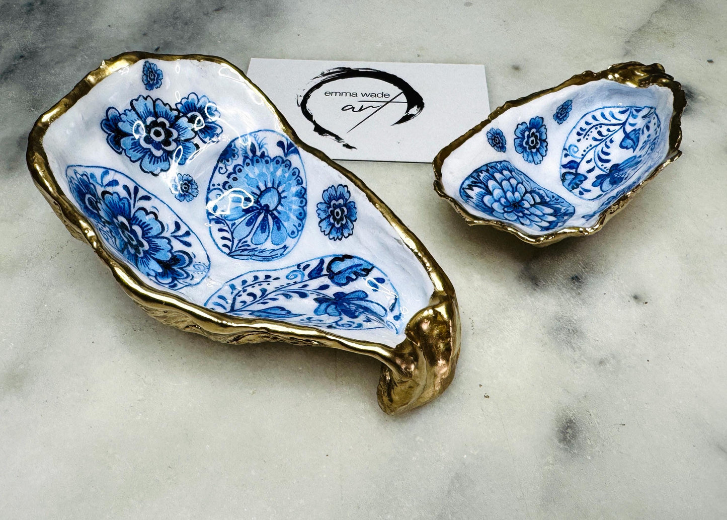 Handmade Dorset oyster shell trinket dish featuring a vintage blue and white decorative egg design with intricate floral porcelain style detailing. Oyster shell is hand gilded with liquid gold leaf edging and sealed in glossy crystal clear resin. Elegant coastal jewelry holder, ring dish, trinket tray, or decorative keepsake inspired by antique ceramics and vintage decor styling. Luxury handmade shell art photographed on a marble background.