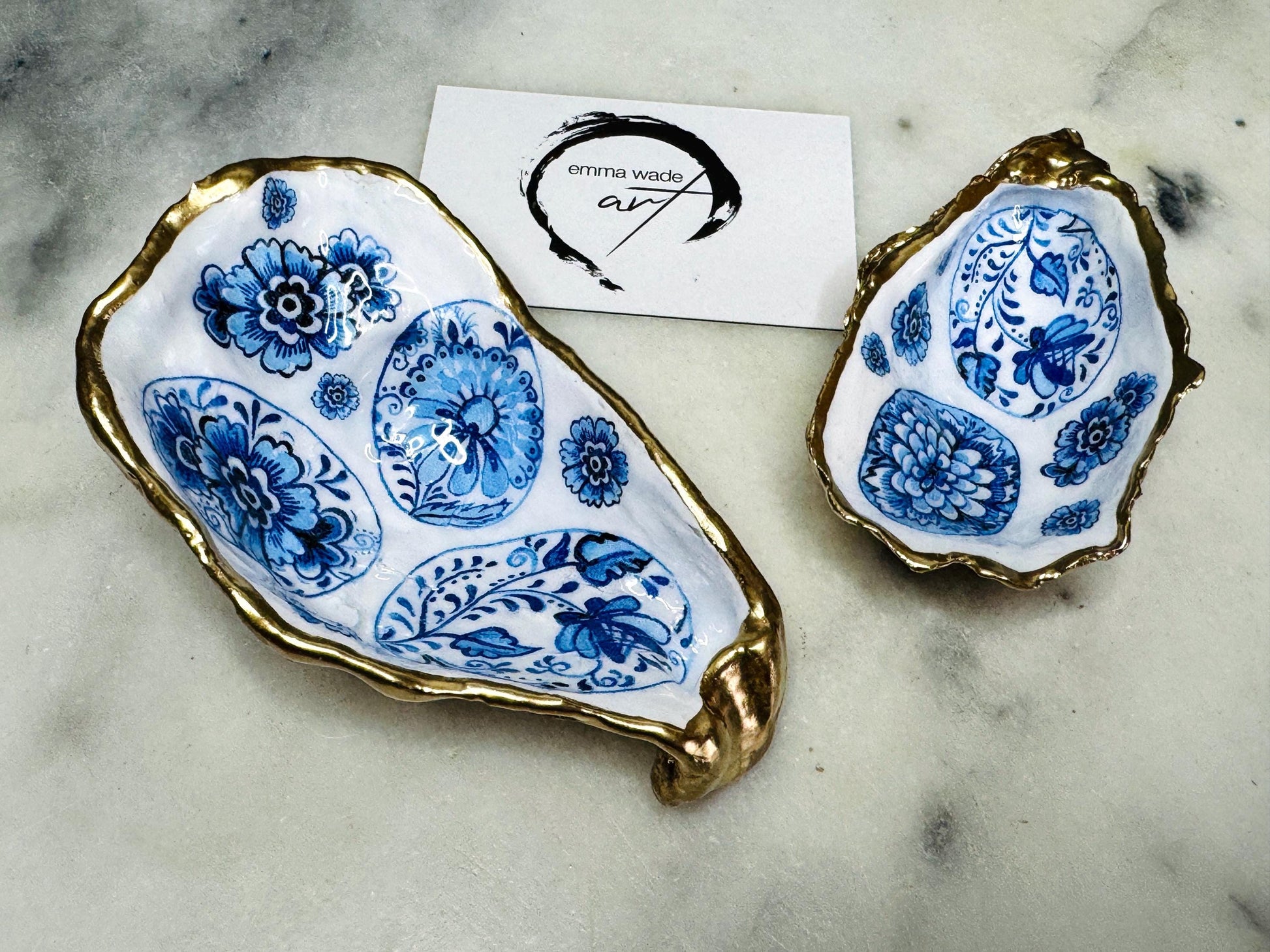 Handmade Dorset oyster shell trinket dish featuring a vintage blue and white decorative egg design with intricate floral porcelain style detailing. Oyster shell is hand gilded with liquid gold leaf edging and sealed in glossy crystal clear resin. Elegant coastal jewelry holder, ring dish, trinket tray, or decorative keepsake inspired by antique ceramics and vintage decor styling. Luxury handmade shell art photographed on a marble background.