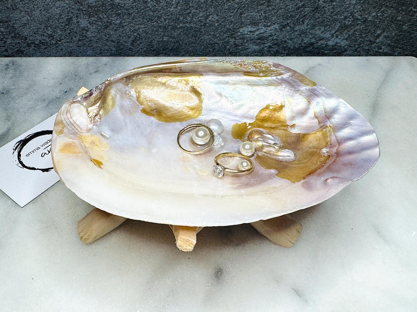 Handmade coastal trinket dish made from a natural iridescent oyster shell with gold detailing and Swarovski pearls acting as ring rests. Displayed on a driftwood style stand, this elegant jewellery holder is perfect as a ring tray, vanity dish, eco friendly gift, beach wedding accessory, coastal home décor piece, or unique artisan keepsake. Ideal for rings, earrings, and small treasures.