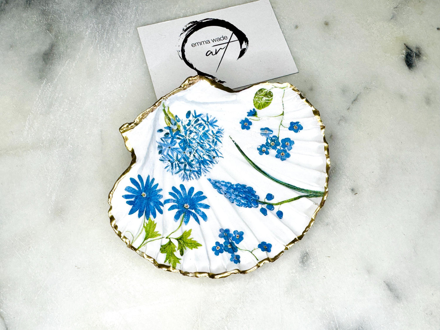 Handcrafted Dorset scallop shell trinket dish with blue spring flower design, finished with a gold leaf rim and glossy resin. The shell shows delicate blue floral motifs with green stems on a natural shell, styled on a light background. A coastal inspired jewellery or ring holder, perfect as a decorative keepsake dish, bathroom accent, or thoughtful handmade gift.