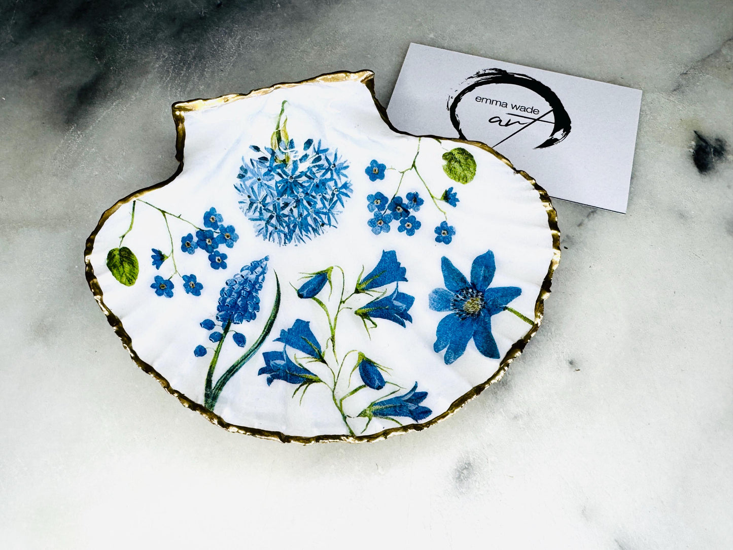 Handcrafted Dorset scallop shell trinket dish with blue spring flower design, finished with a gold leaf rim and glossy resin. The shell shows delicate blue floral motifs with green stems on a natural shell, styled on a light background. A coastal inspired jewellery or ring holder, perfect as a decorative keepsake dish, bathroom accent, or thoughtful handmade gift.