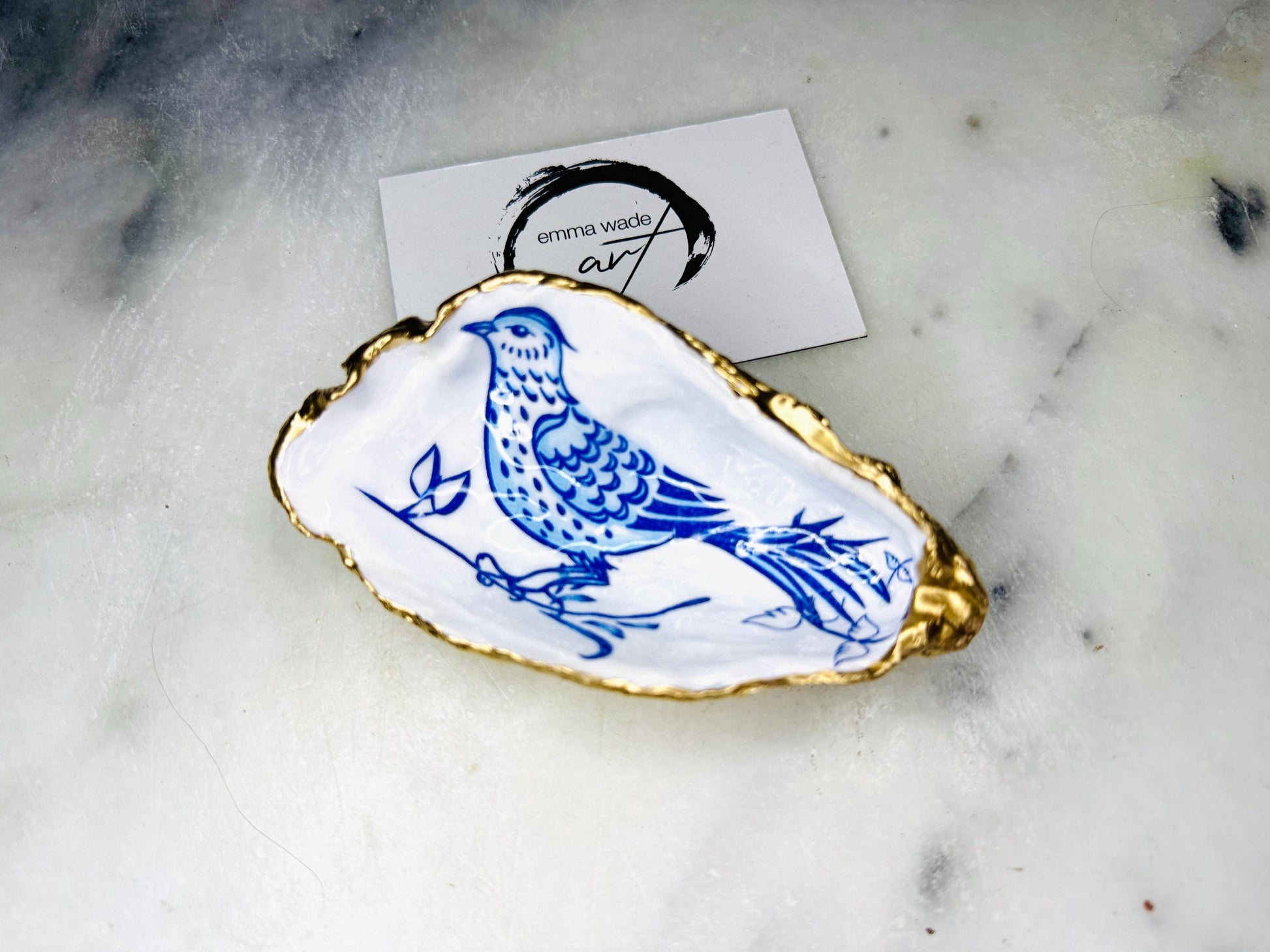 Handmade Dorset oyster shell trinket dish with blue porcelain-style bird design, gold leaf gilded edges and glossy resin finish. Coastal jewellery holder and ring dish photographed on a light surface, showing the natural shell shape and pearly interior. Unique sustainably sourced shell, sold individually as decorative coastal home décor or a thoughtful gift.