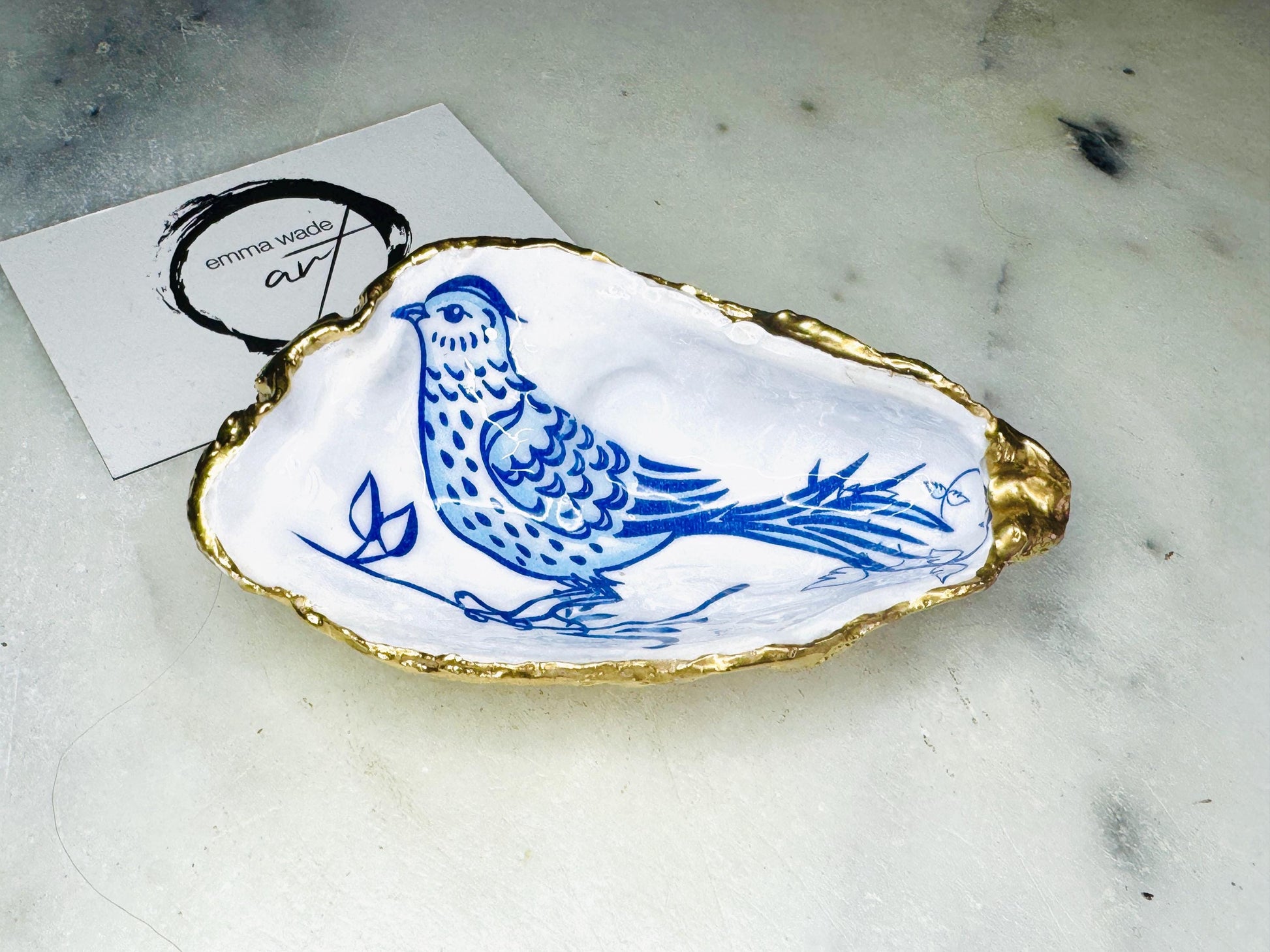 Handmade Dorset oyster shell trinket dish with blue porcelain-style bird design, gold leaf gilded edges and glossy resin finish. Coastal jewellery holder and ring dish photographed on a light surface, showing the natural shell shape and pearly interior. Unique sustainably sourced shell, sold individually as decorative coastal home décor or a thoughtful gift.