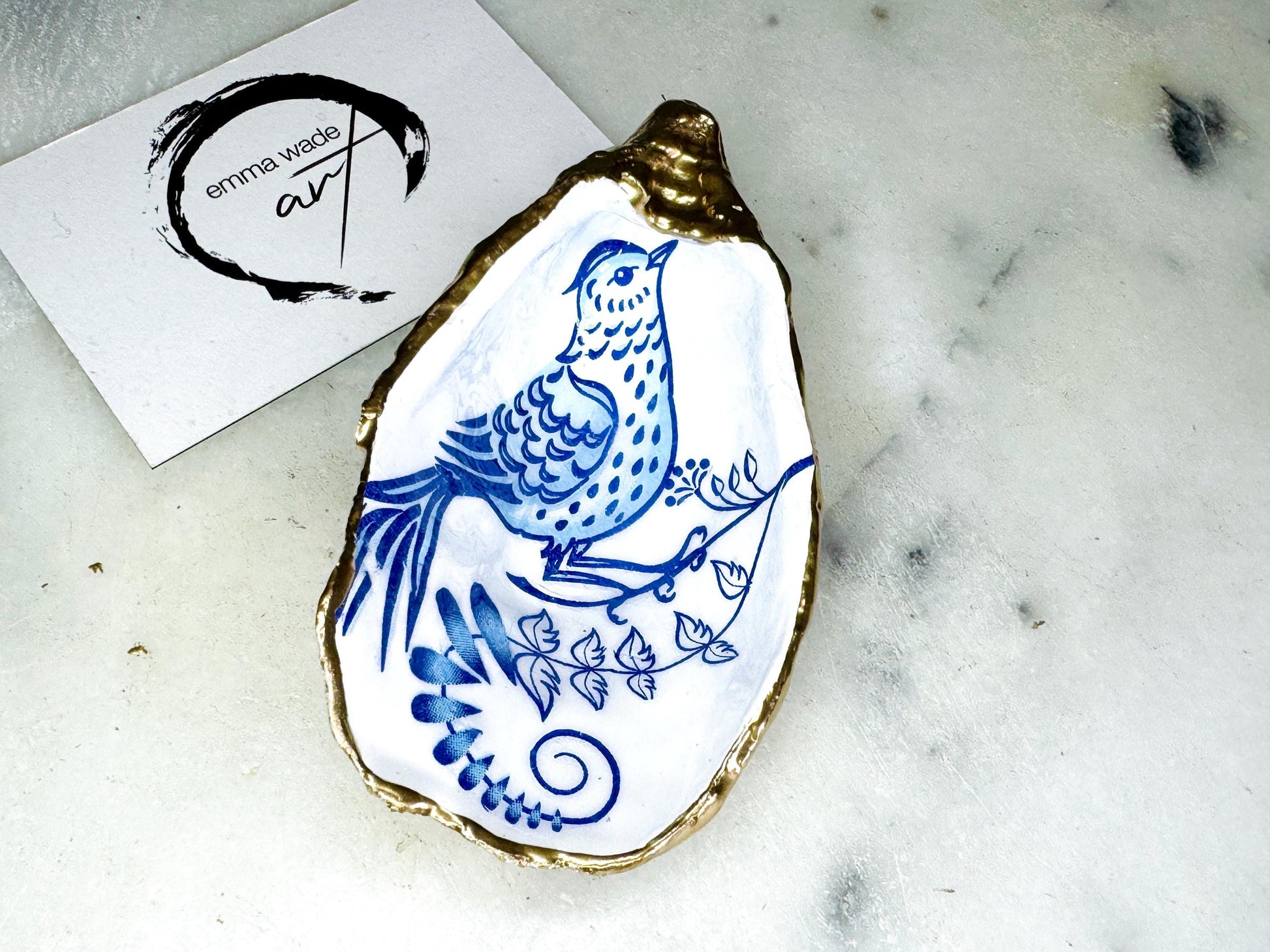 Handmade Dorset oyster shell trinket dish with blue porcelain-style bird design, gold leaf gilded edges and glossy resin finish. Coastal jewellery holder and ring dish photographed on a light surface, showing the natural shell shape and pearly interior. Unique sustainably sourced shell, sold individually as decorative coastal home décor or a thoughtful gift.